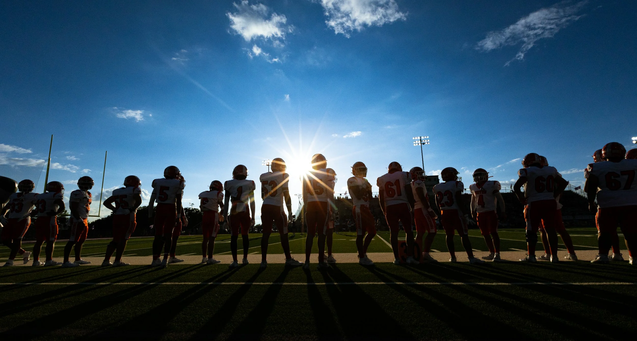 Silhouettes of football players standing on a field during sunset, with sun behind them and shadows cast on the ground.