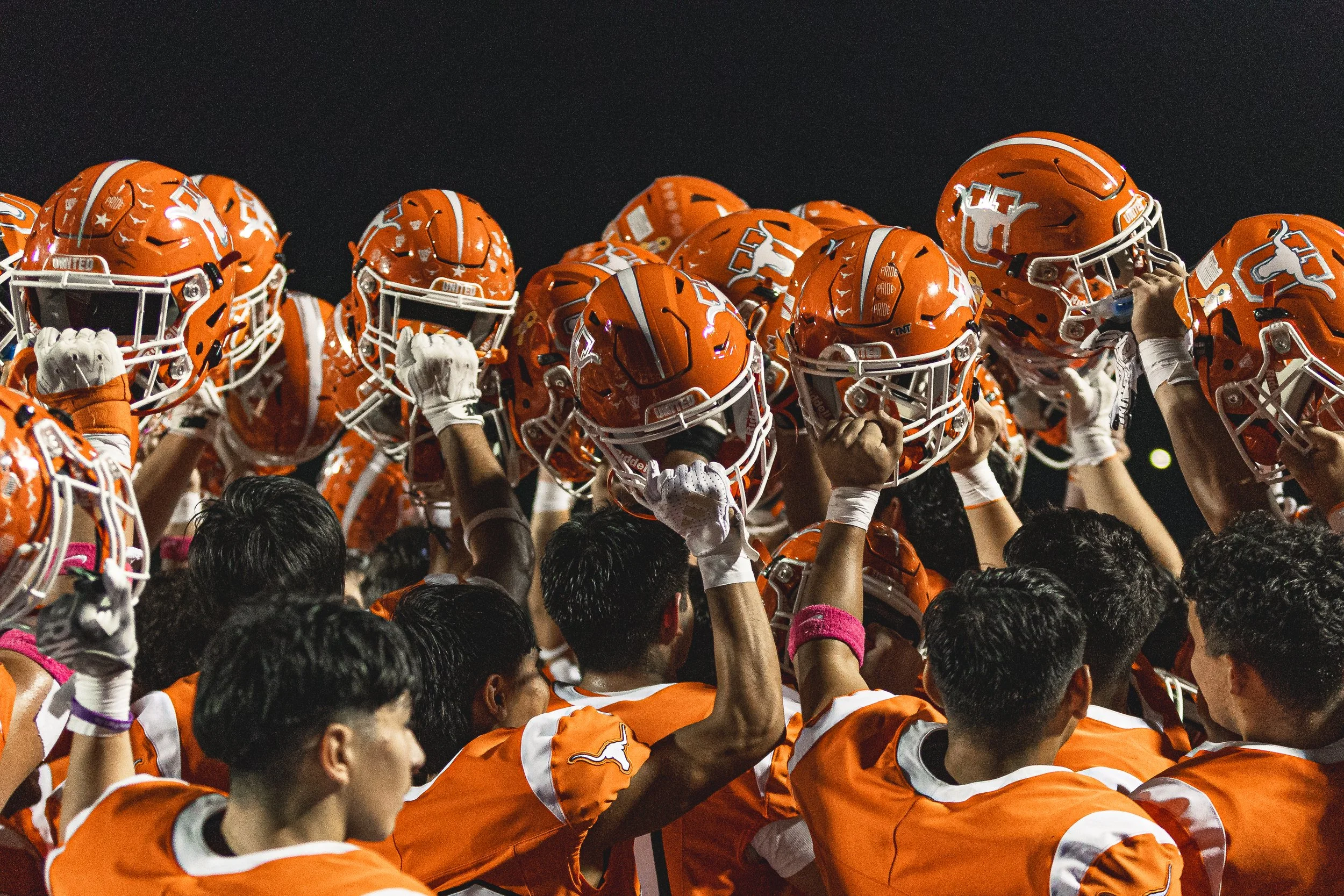 A football team wearing orange uniforms holding their helmets in the air during a night game.