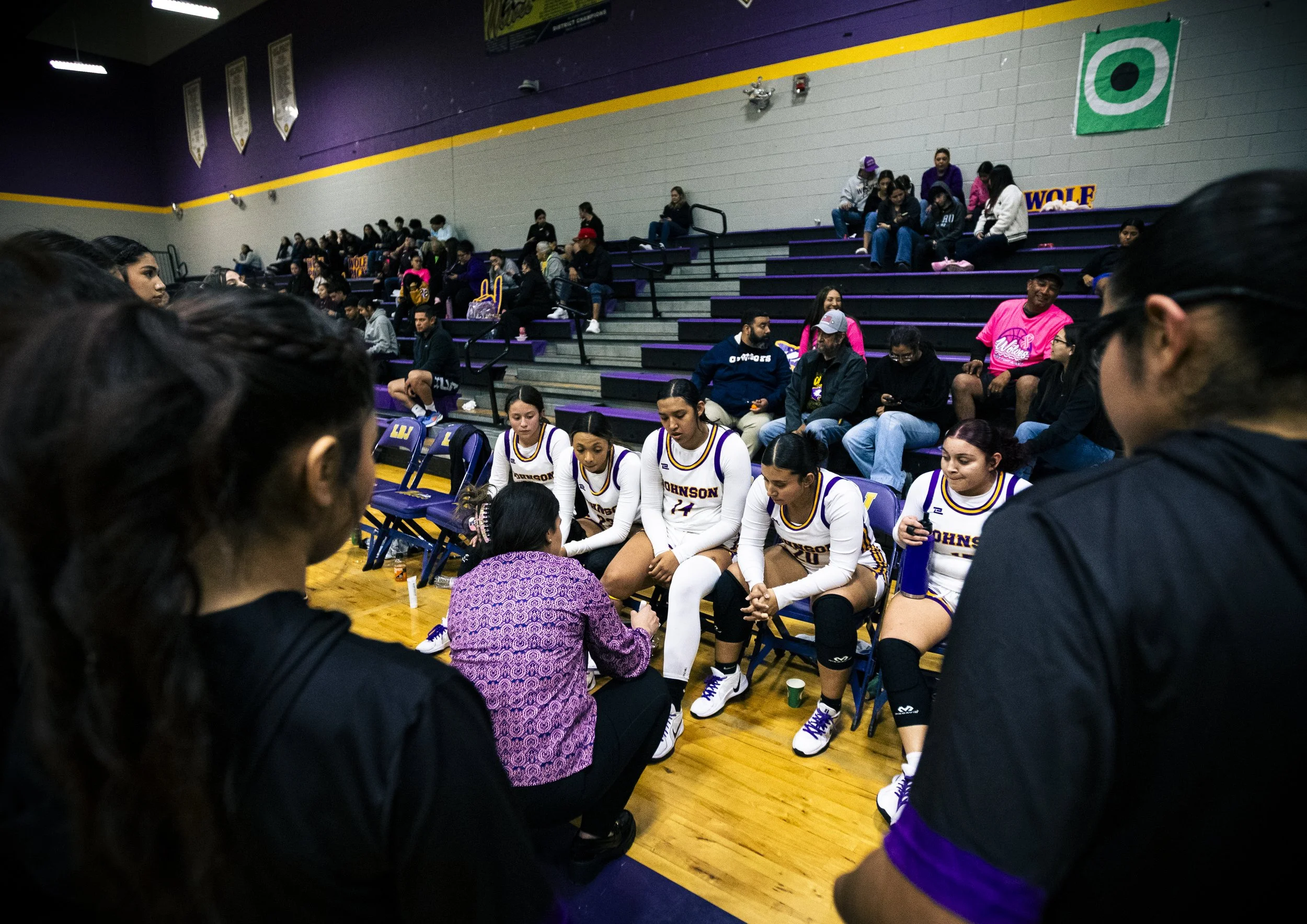 Girls' basketball team sits on bench while coach speaks to them during game in gymnasium with spectators on bleachers in background.