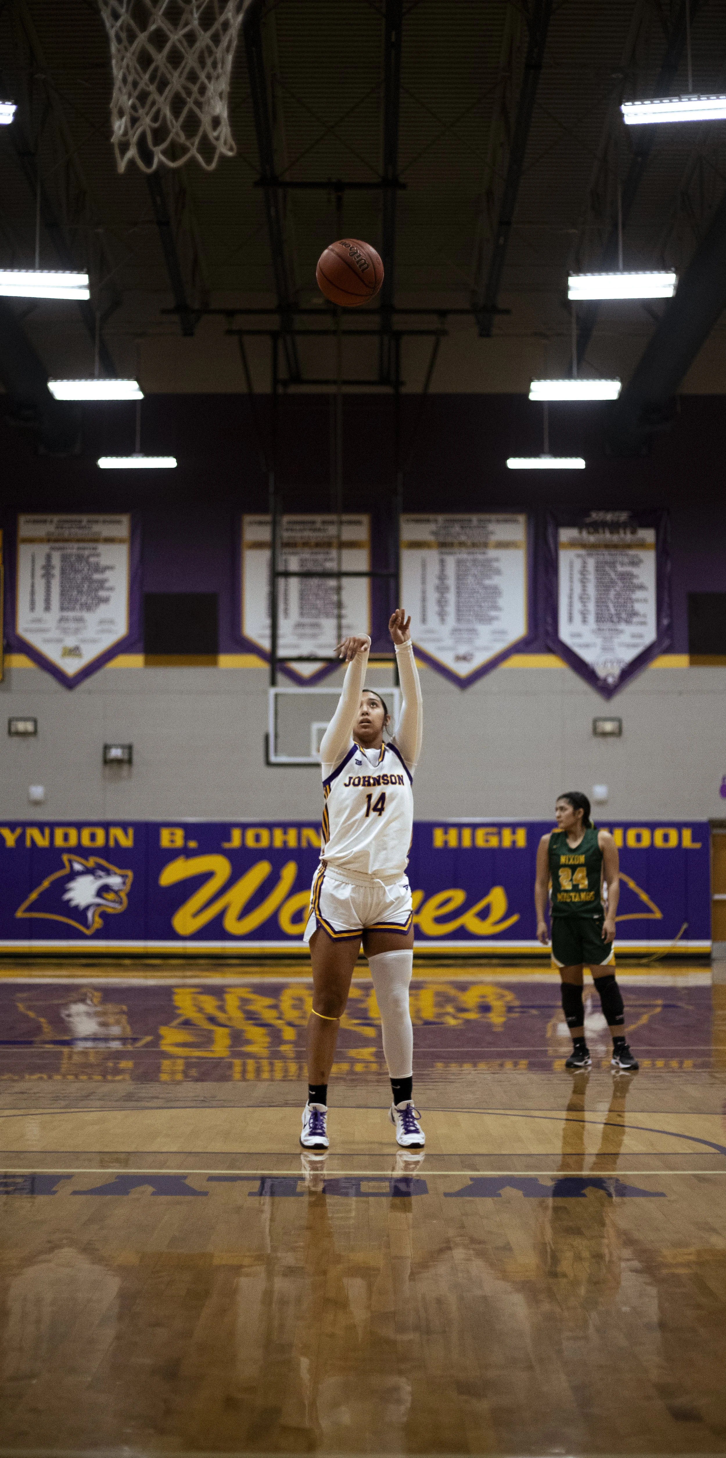 A female basketball player in a whites and purple uniform, number 14, is shooting a free throw in an indoor basketball court. Another player in green and yellow stands in the background. The court features purple and yellow branding for Lyndon B. Joh