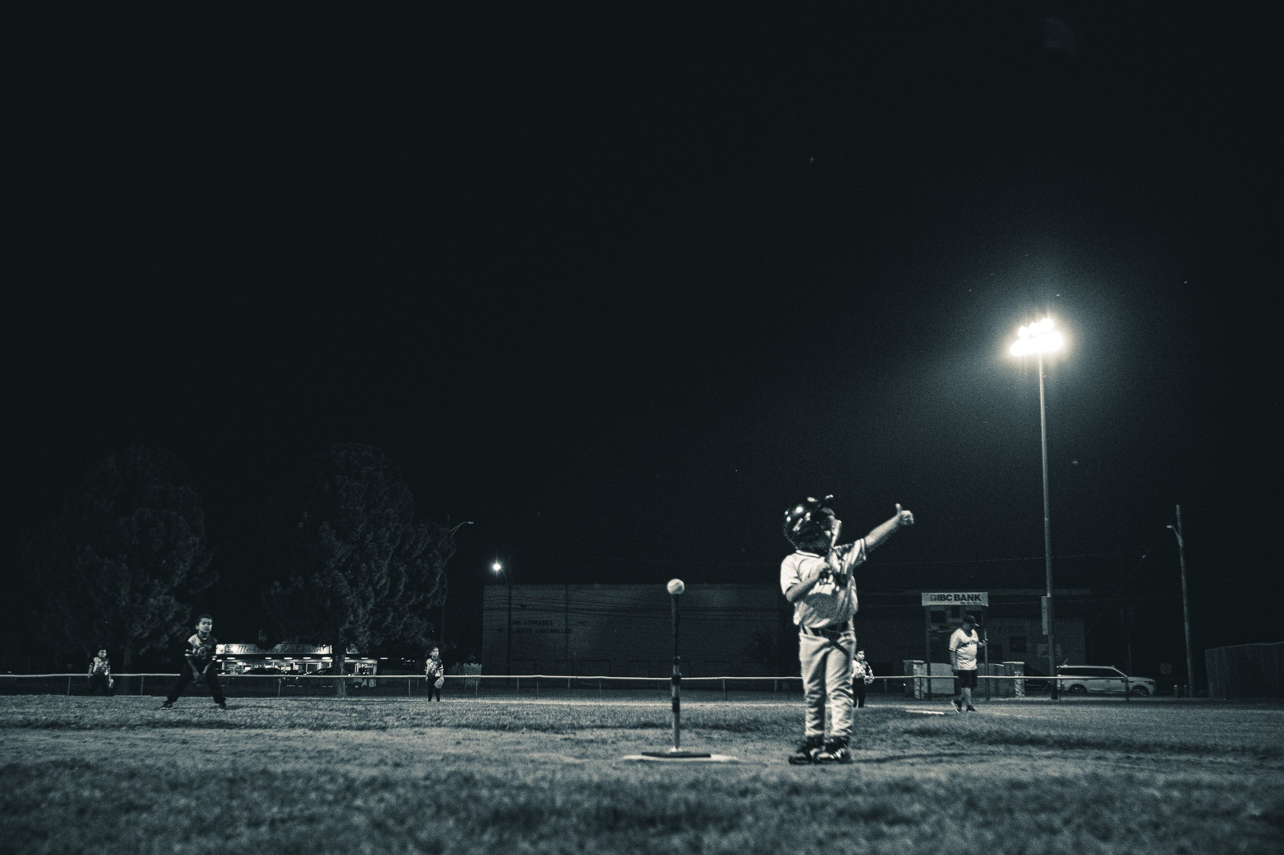 Nighttime scene of children playing baseball on a field, with one child at the batter's box, and others in the field, under bright floodlights.