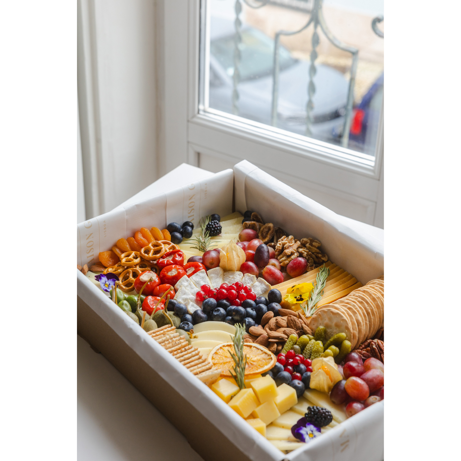 A cheese and fruit platter with grapes, berries, nuts, cheese, crackers, and dried fruit, set near a window.