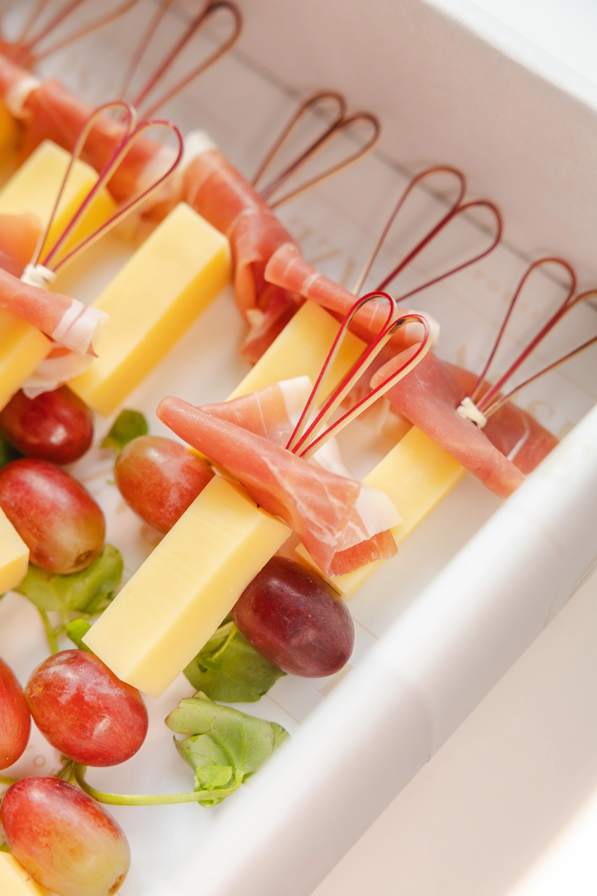 Cheese and grape charcuterie arrangement with slices of prosciutto and skewers on a white tray.