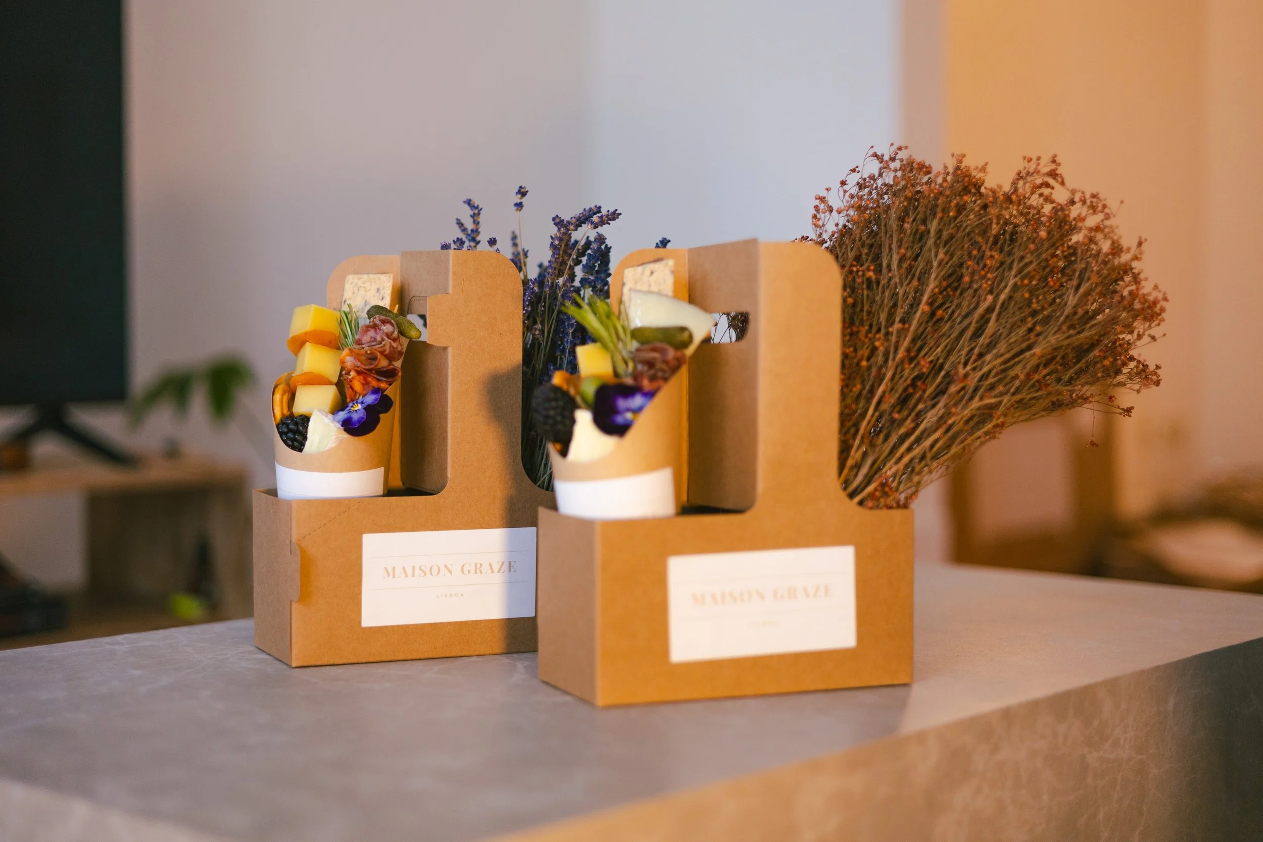 Two cardboard boxes labeled 'MAISON GRAZE' contain flower arrangements with dried and fresh flowers, arranged on a table in warm indoor lighting.