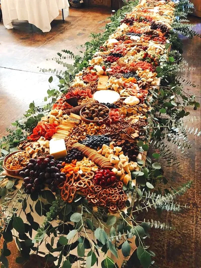 Long table decorated with greenery, filled with assorted cheeses, crackers, grapes, berries, and charcuterie.