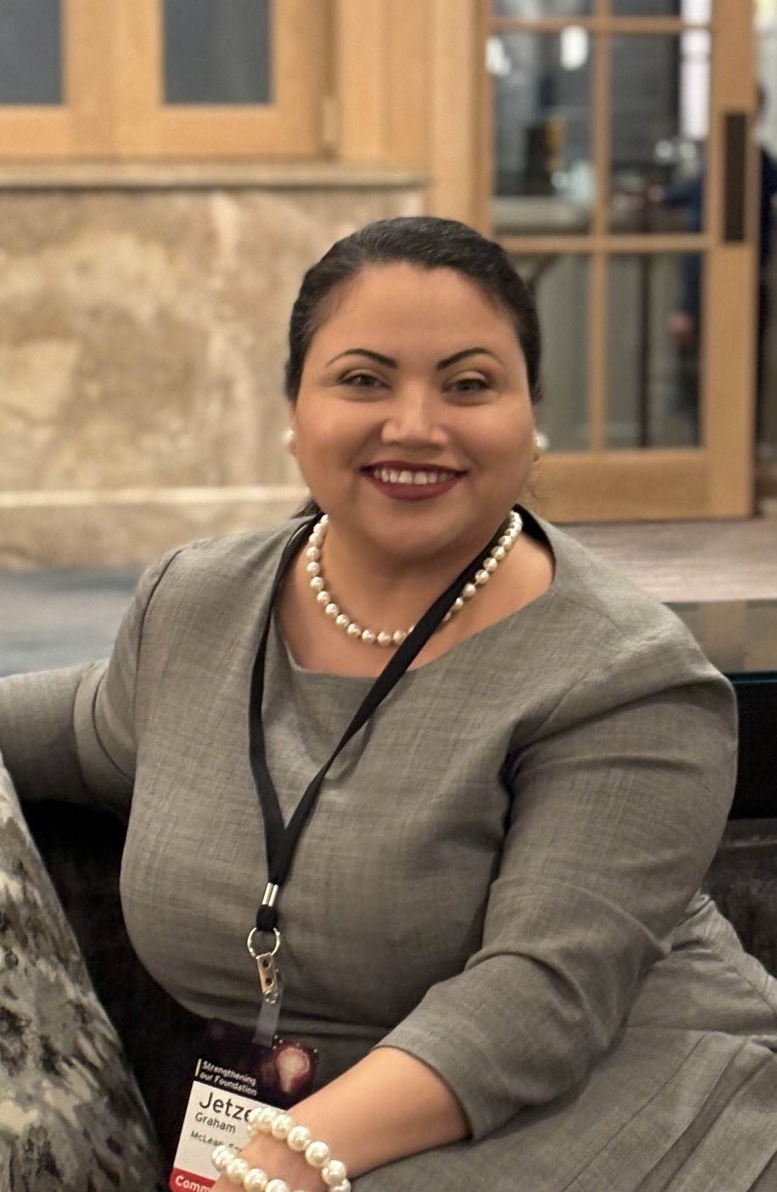 A woman with dark hair in a bun, wearing a grey suit, pearl necklace, pearl earrings, and a bracelet, smiling while sitting at an indoor event with a wooden wall and glass window in the background.