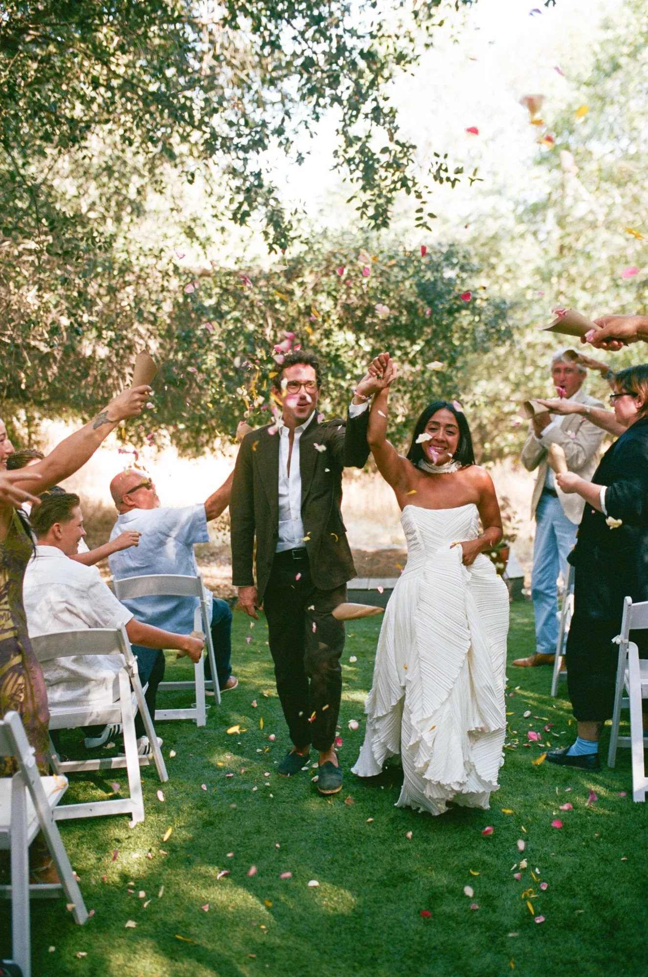 A newlywed couple walking hand in hand through a garden aisle while guests throw flower petals, celebrating their wedding.