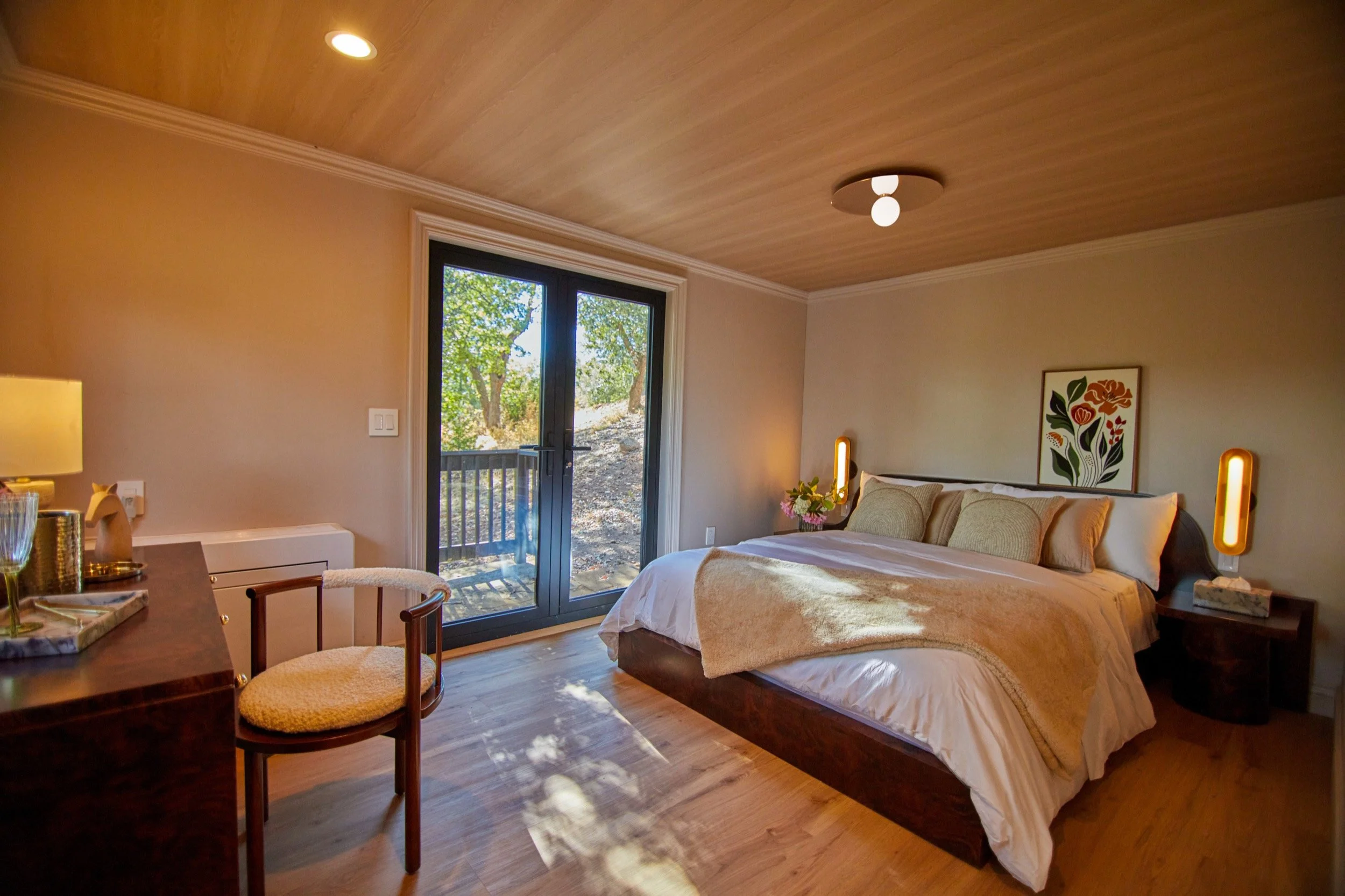 A cozy bedroom with a large bed, beige bedding, and pillows, next to a window with a scenic view of trees, decorated with modern art and warm lighting.