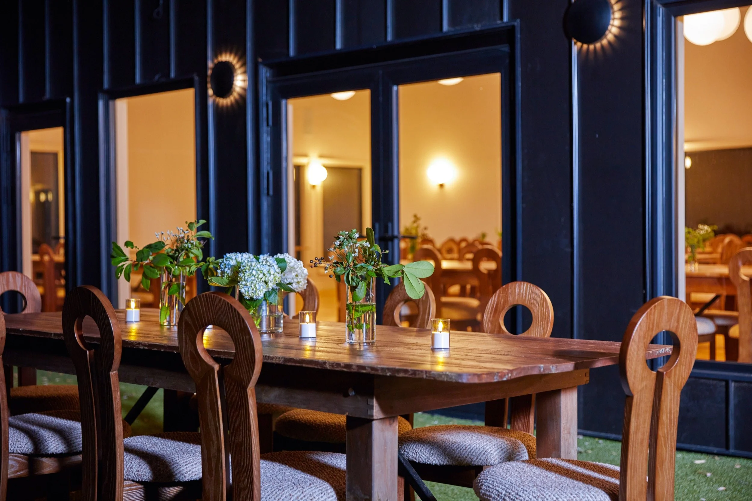 A dinner table set with white hydrangeas and candles outside tall glass doors at dusk.