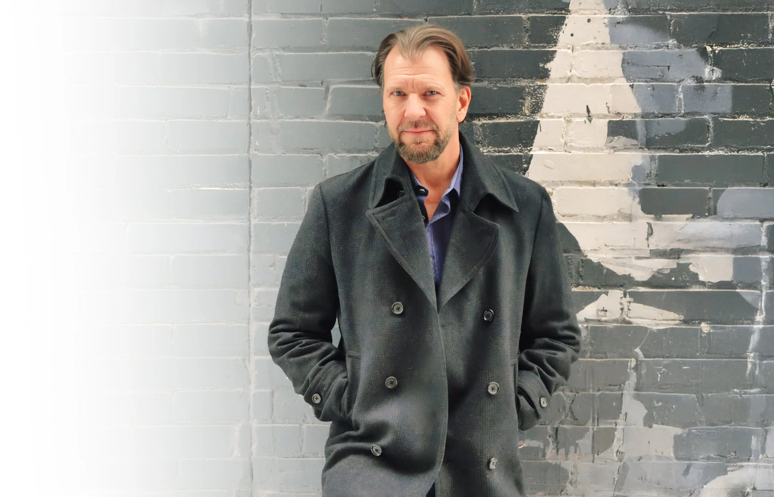 A middle-aged man with brown hair and a beard standing in front of a brick wall with a mural. He is wearing a dark gray peacoat and has his hands in his coat pockets, looking at the camera with a neutral expression.