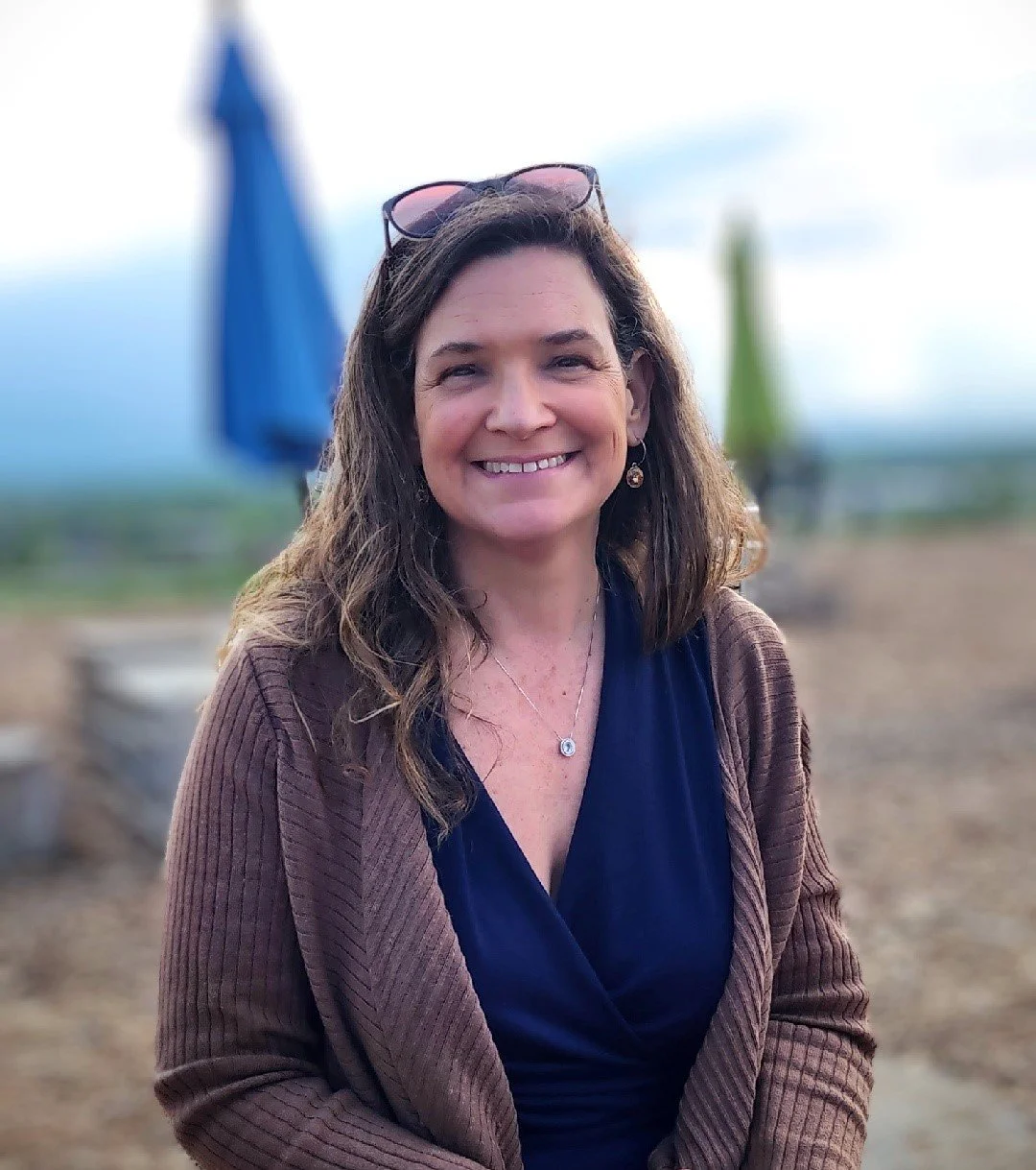 A woman with brown hair and sunglasses on her head, smiling, outdoors with umbrellas in the background.