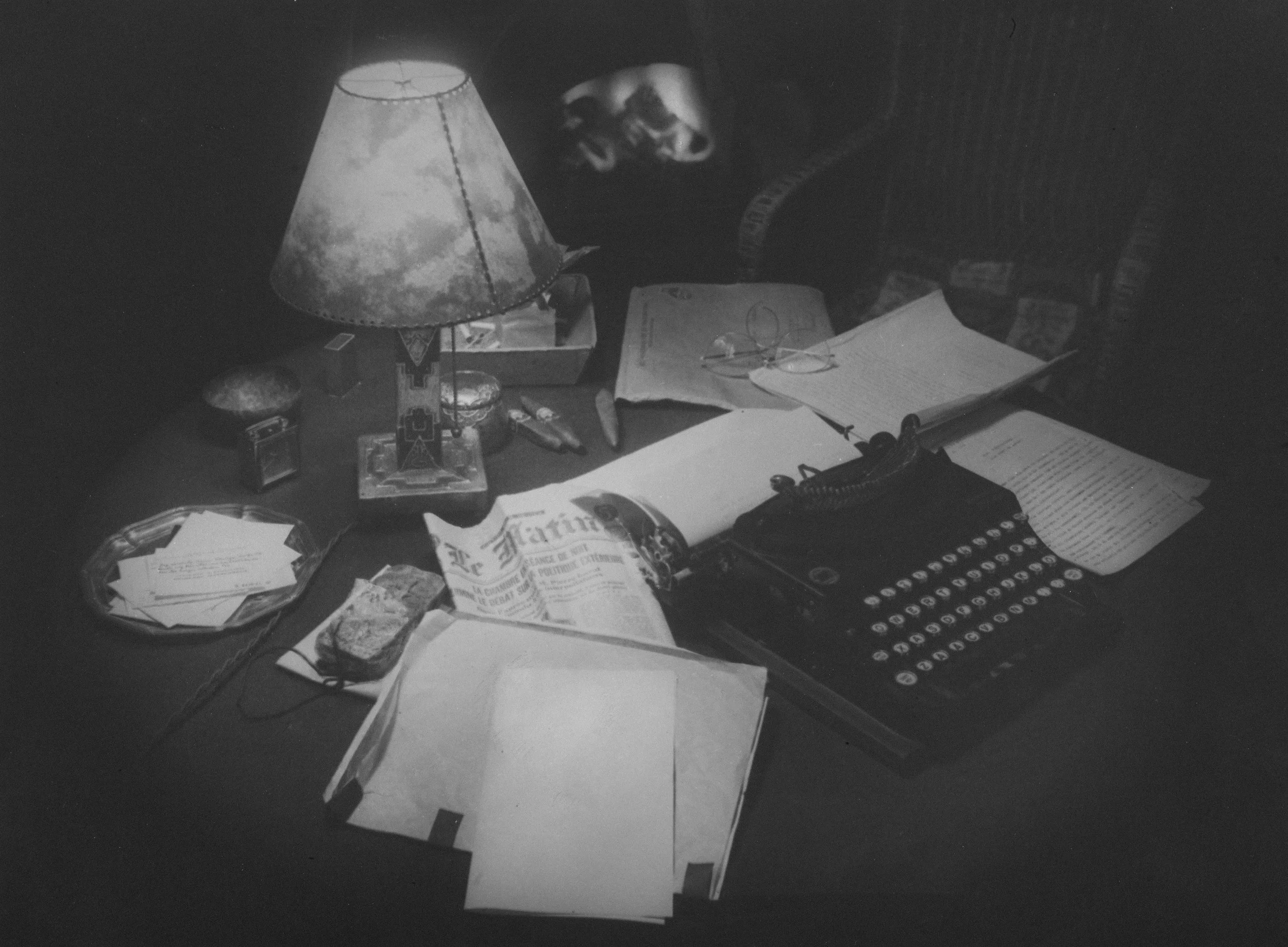 A cluttered desk with a classic typewriter, open books, papers, a desk lamp, and various small objects in black and white.