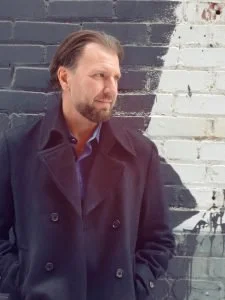 A man with light brown hair and a beard, wearing a dark coat, standing against a painted brick wall with black and white sections.