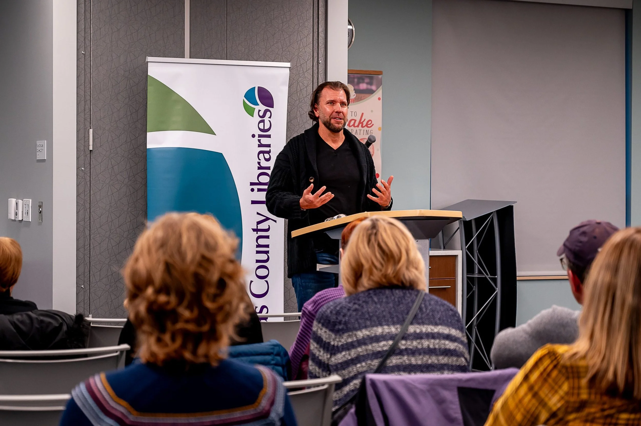 A man giving a speech at a lectern in front of an audience, with a County Libraries banner behind him.