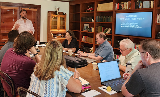 A group of seven people in a meeting room listening to a man standing and speaking. The room has a large wooden bookshelf, a flat-screen TV displaying a presentation, and a wooden door. The table has laptops, notebooks, cups, and other meeting materials.