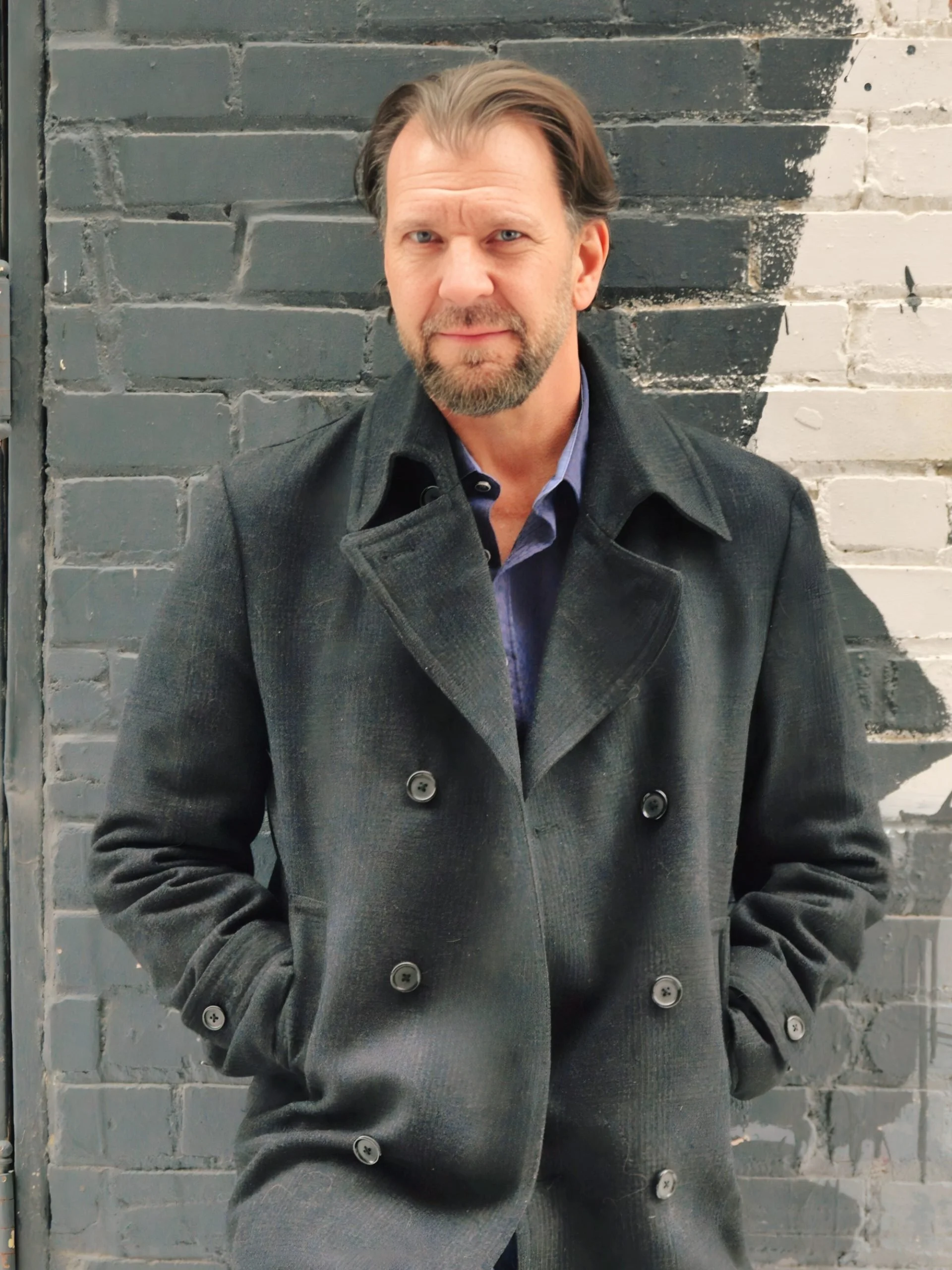Man with brown hair and beard wearing a gray peacoat standing against a gray brick wall with a painted black and white design.