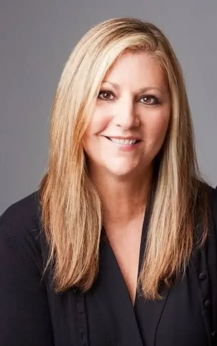 A woman with long blonde hair, smiling, wearing a black top, against a gray background.