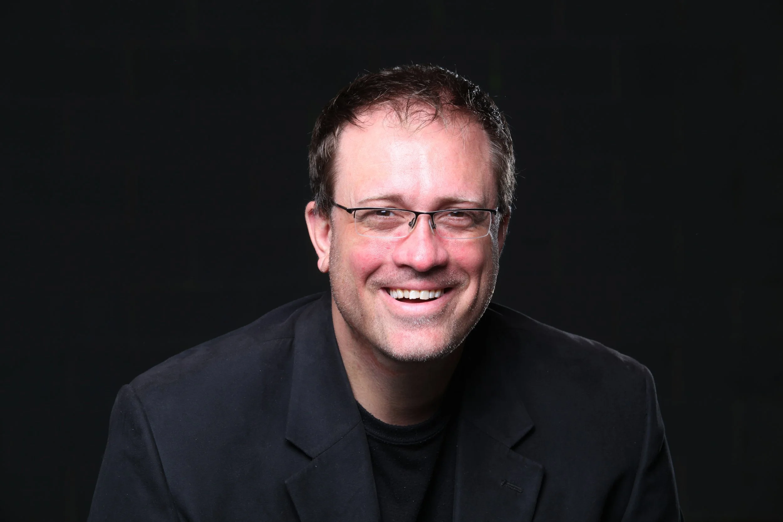 A smiling man with glasses, wearing a black jacket, against a dark background.
