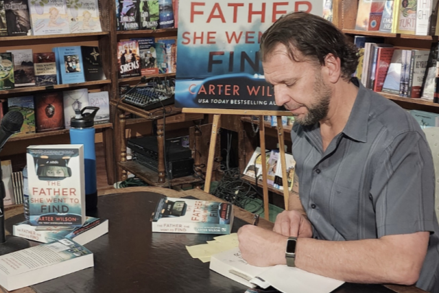Autograph signing event for the book "The Father She Went to Find" by Crter Wilson, featuring a man with dark hair and beard signing a book at a table with promotional materials in a bookstore.