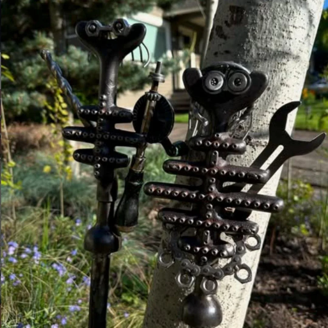 Sculpture made from bike parts, resembling a face with two eyes, a nose, and a mouth, placed on a tree trunk in a garden.
