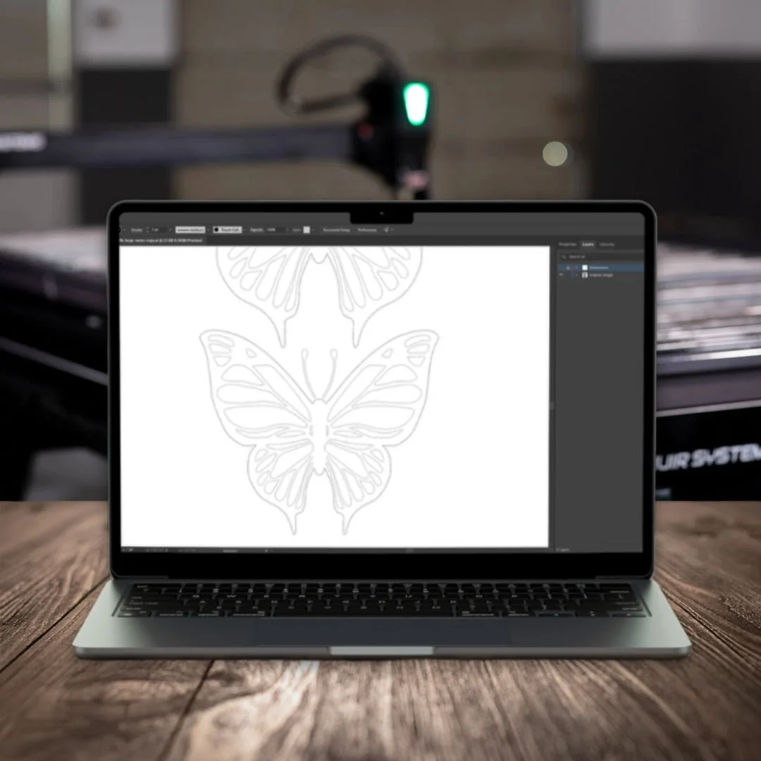 Laptop on a wooden table displaying a butterfly line art design in graphic design software, with a laser cutter in the background.