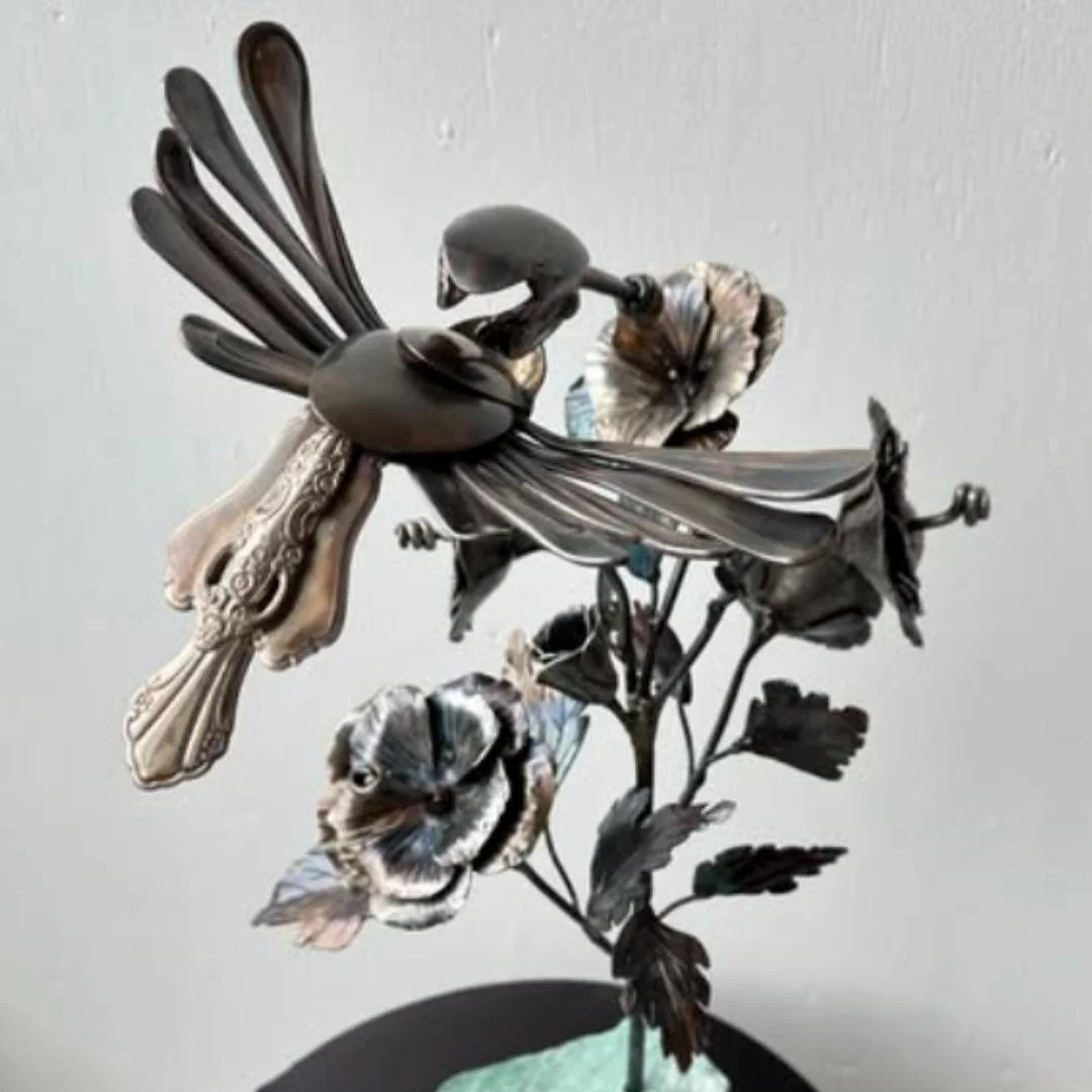 Metal sculpture of a bird perched on a flowering branch against a plain background.