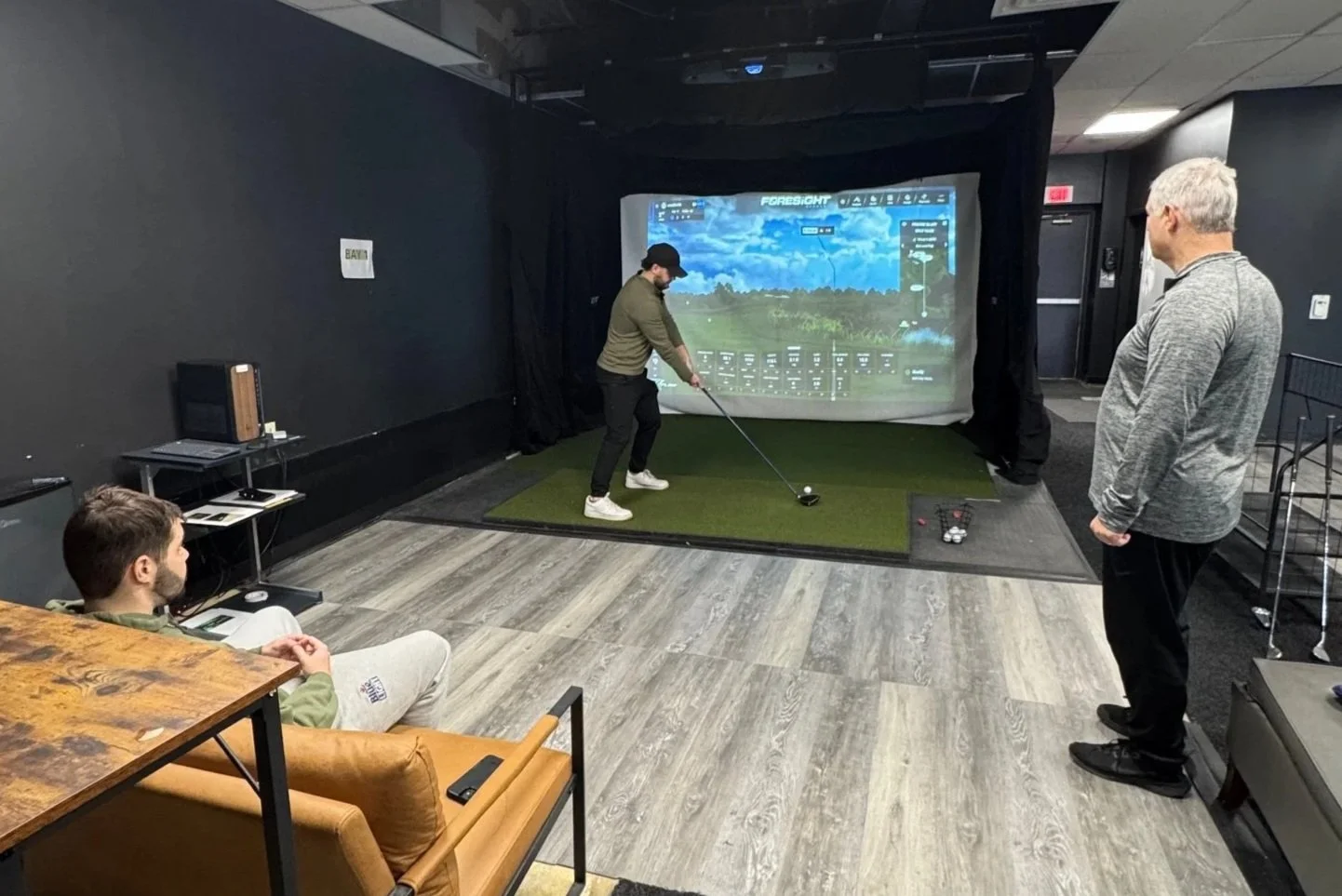 Three men in a room with an indoor golf simulator. One man in a gray jacket watches as another man swings a golf club, aiming at a screen showing a golf course. The third man sits on a couch observing. The room has wooden flooring and a mix of modern and casual furniture.