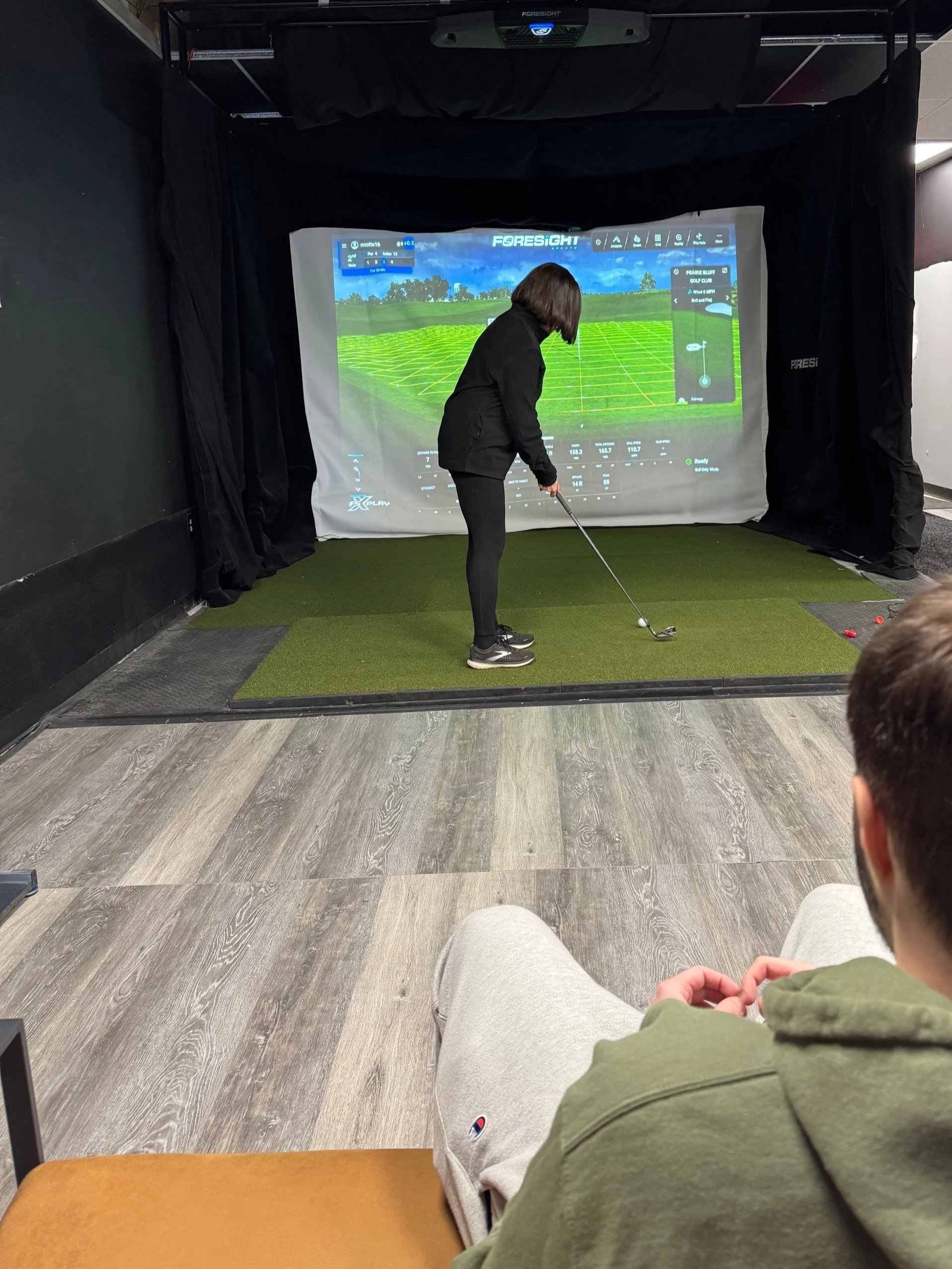 Indoor golf simulator with a woman practicing her swing in front of a large screen showing a virtual golf course, while a man watches from a couch.