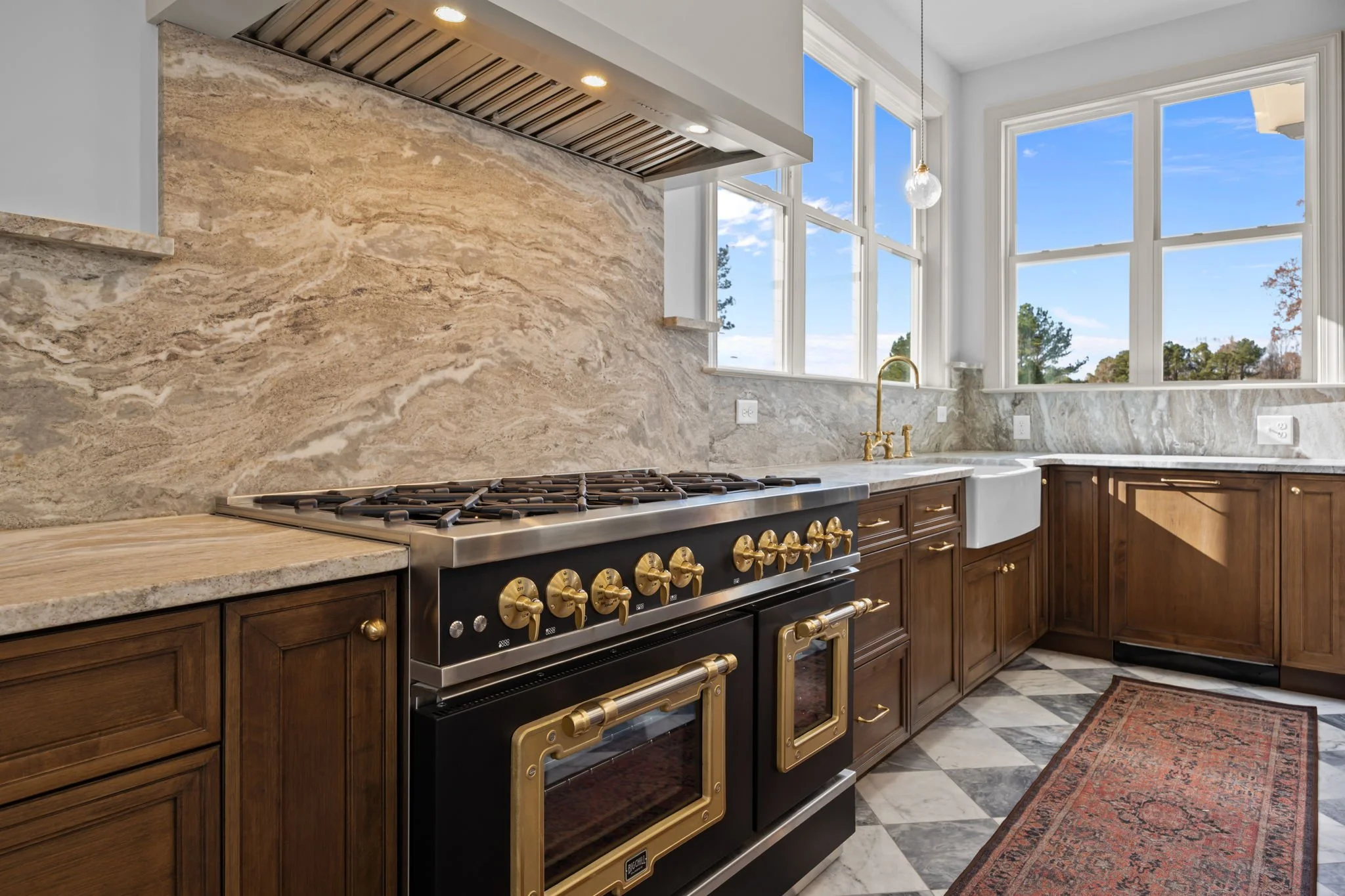 Kitchen with wooden cabinets, a black and gold stove, marble countertops, a white farmhouse sink, large windows showing blue sky and trees, and a patterned rug on a checkered floor.