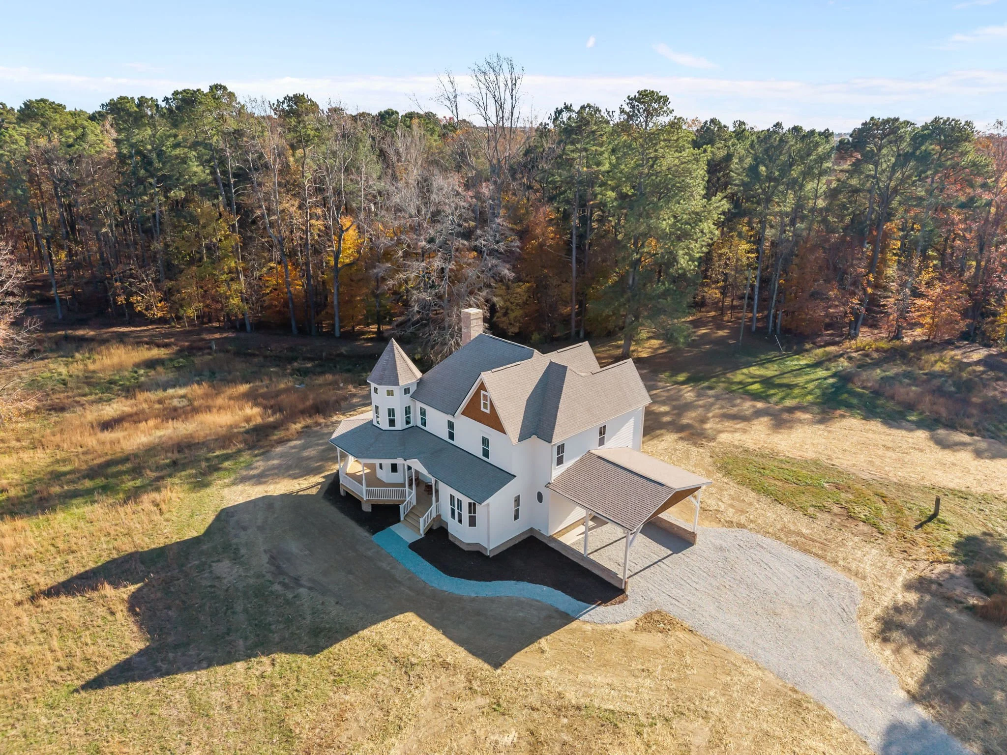 A white house with multiple gabled roofs and a turret, surrounded by a partially cleared yard, with a gravel driveway and wooded area in the background.