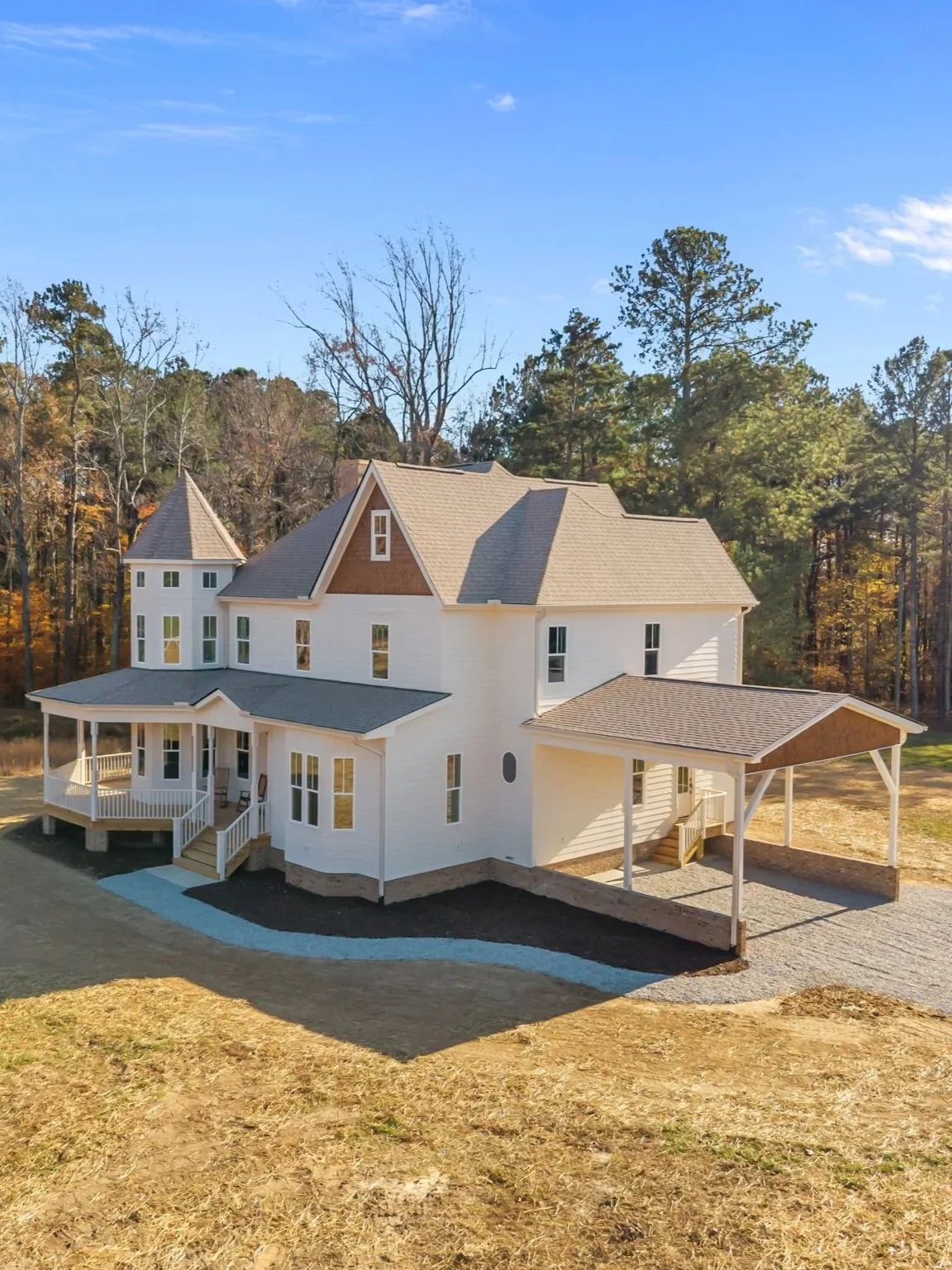 A newly built white house with multiple gabled roofs, a small front porch, and a driveway. The house is situated in a wooded area with trees in the background.