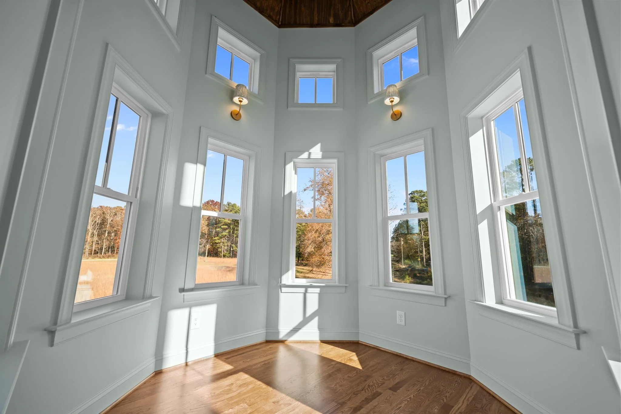 Empty sunroom with multiple windows, white walls, hardwood flooring, and wall-mounted lamps showing a view of the outdoors with trees and a blue sky.