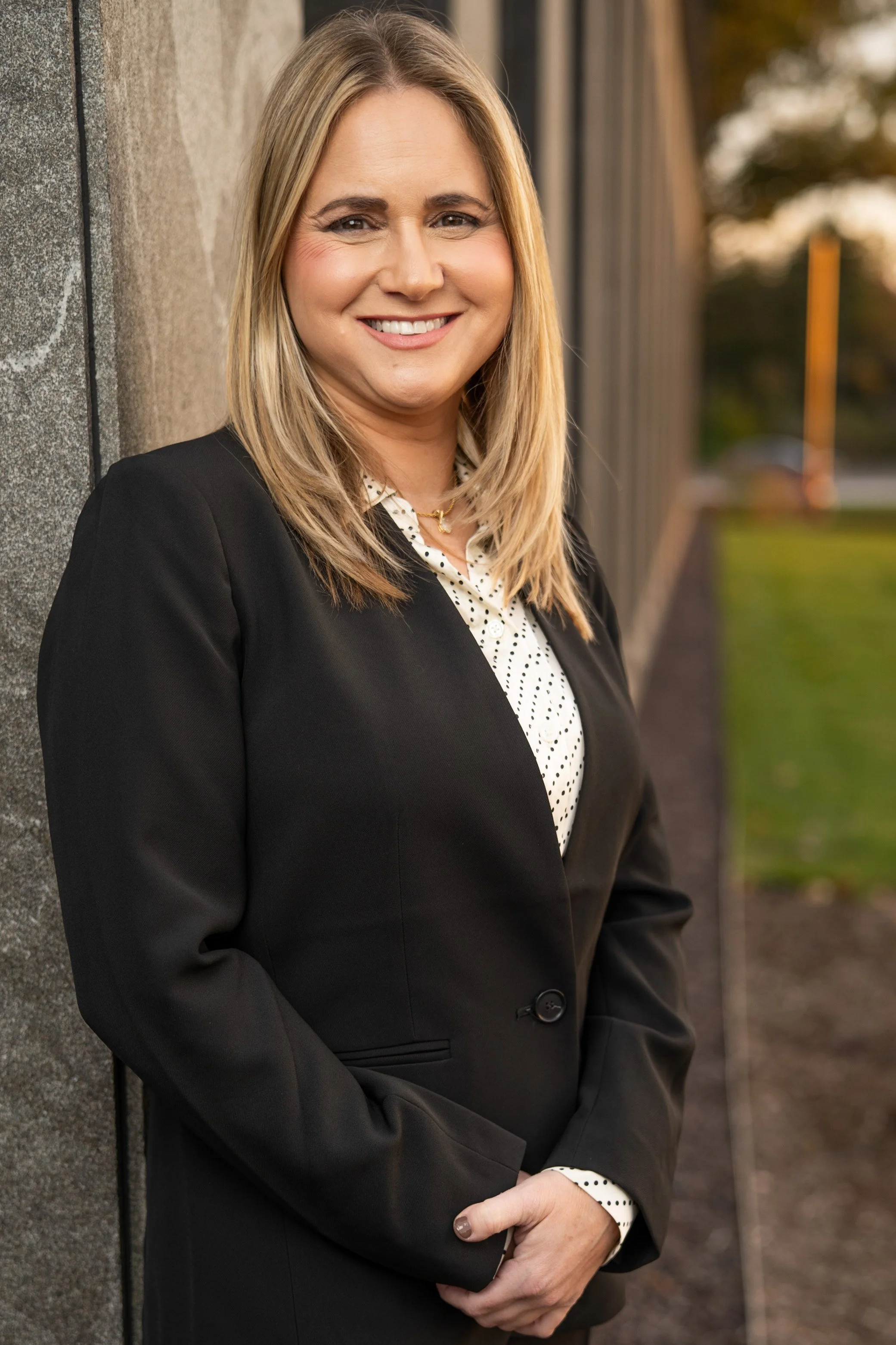 A professional woman with shoulder-length blonde hair, dressed in a black blazer and a white blouse with black polka dots, smiling while standing outdoors against a wall during sunset.