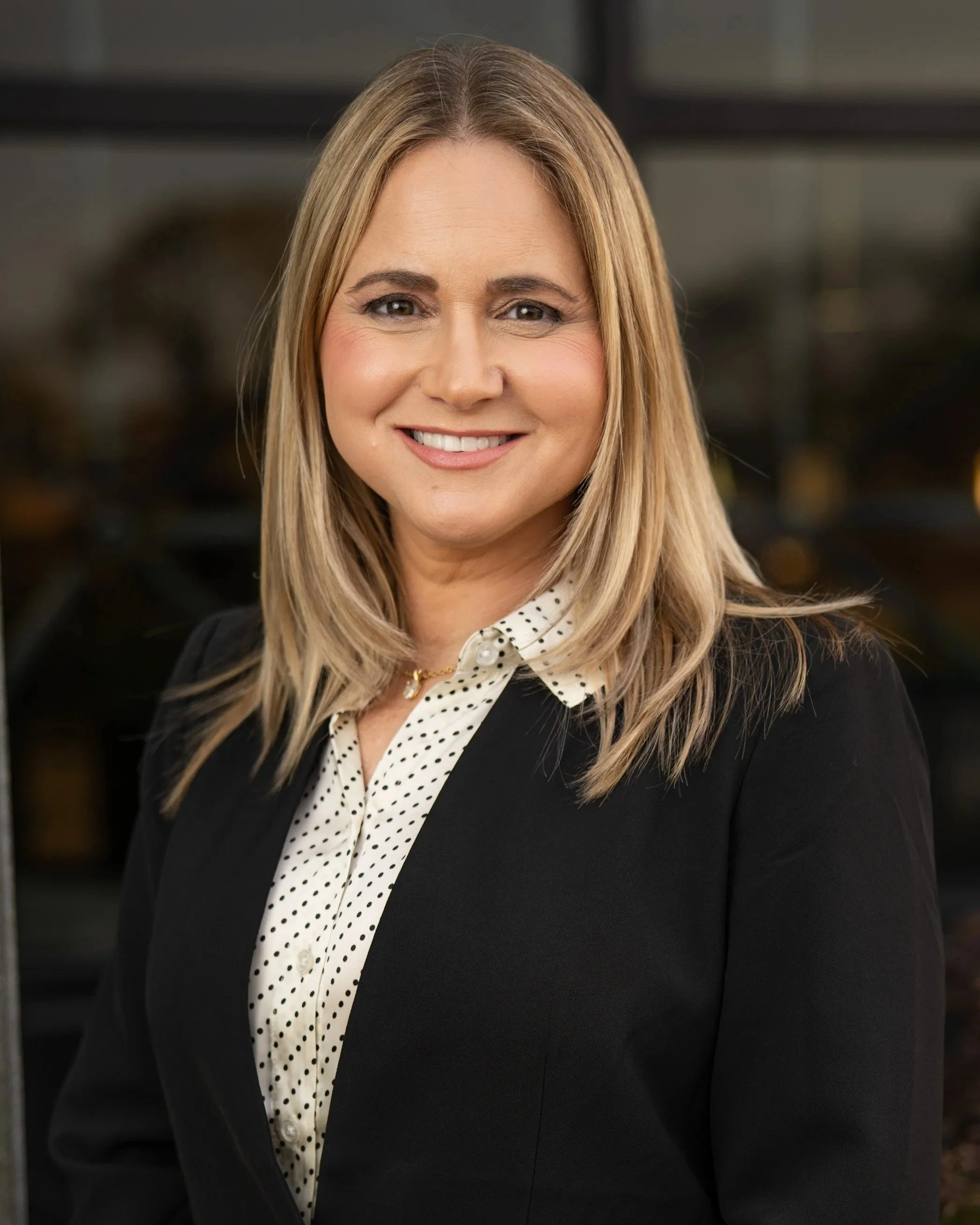 A professional woman with shoulder-length blonde hair, smiling, dressed in a black blazer and a white blouse with black polka dots standing in front of a building.