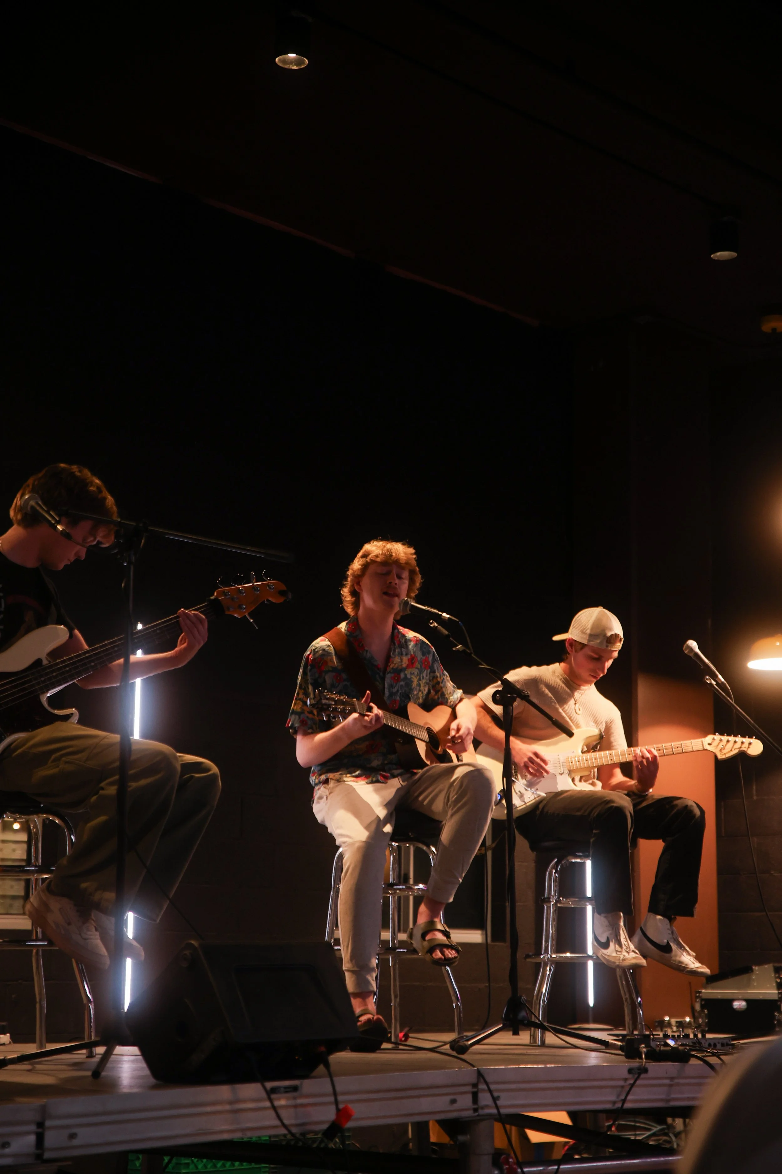 Three young men perform on stage with musical instruments, one singing into a microphone, with a dark background and stage lighting.