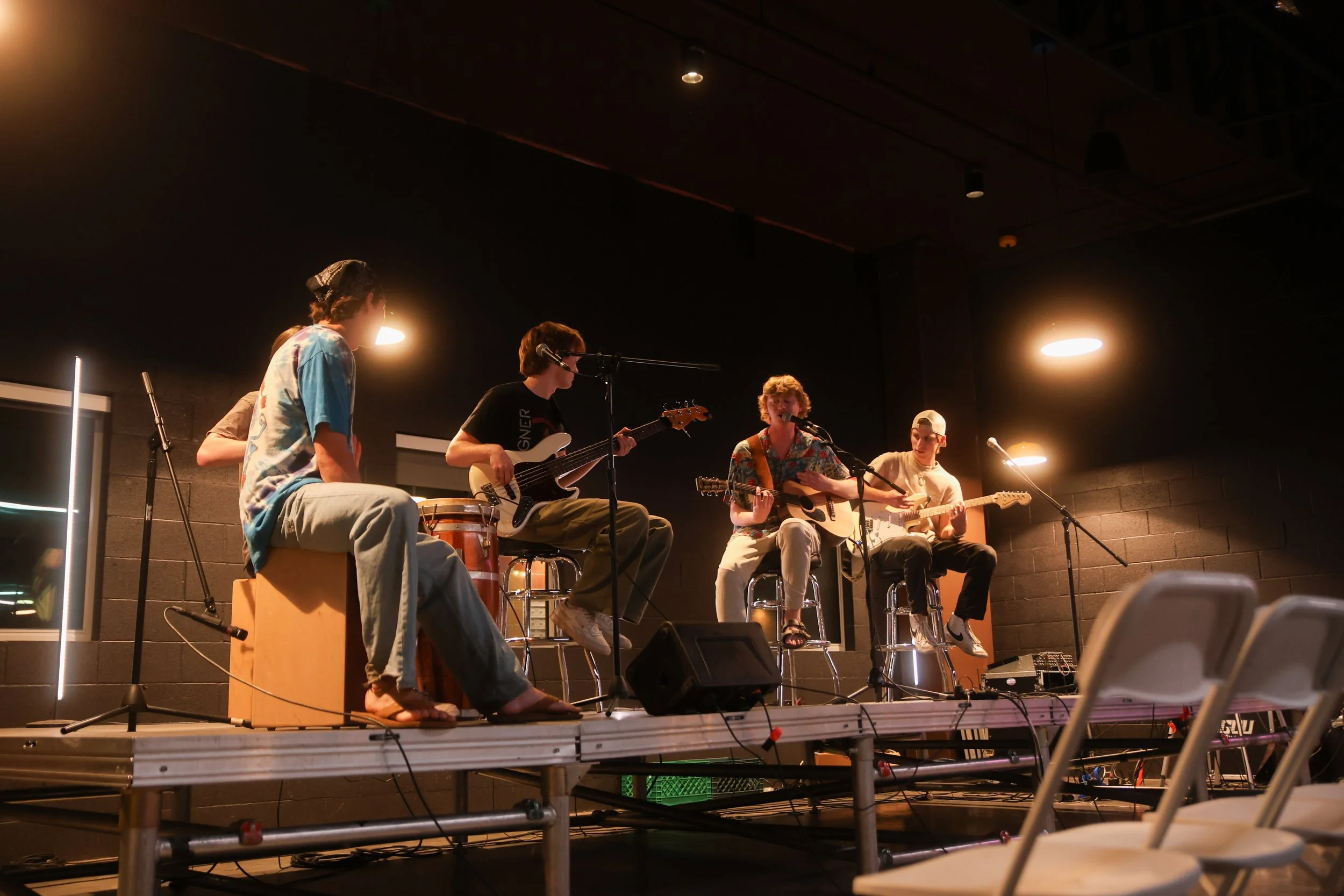 A band of five young men performing on a small stage with musical instruments including guitars, a bass, and percussion, in a dimly lit indoor venue.