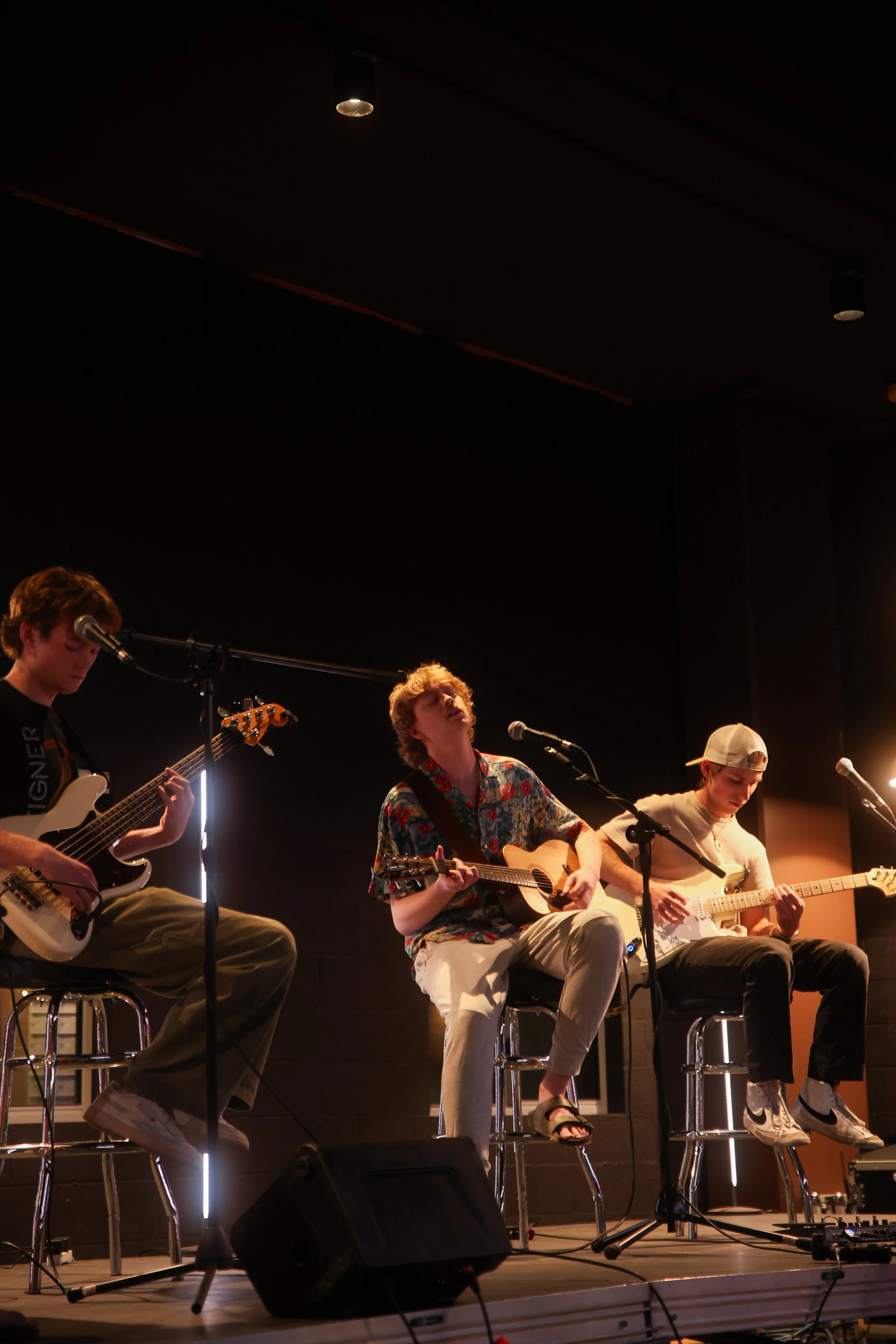 Three young men playing guitars on a stage during a live performance, with microphones and stage equipment.