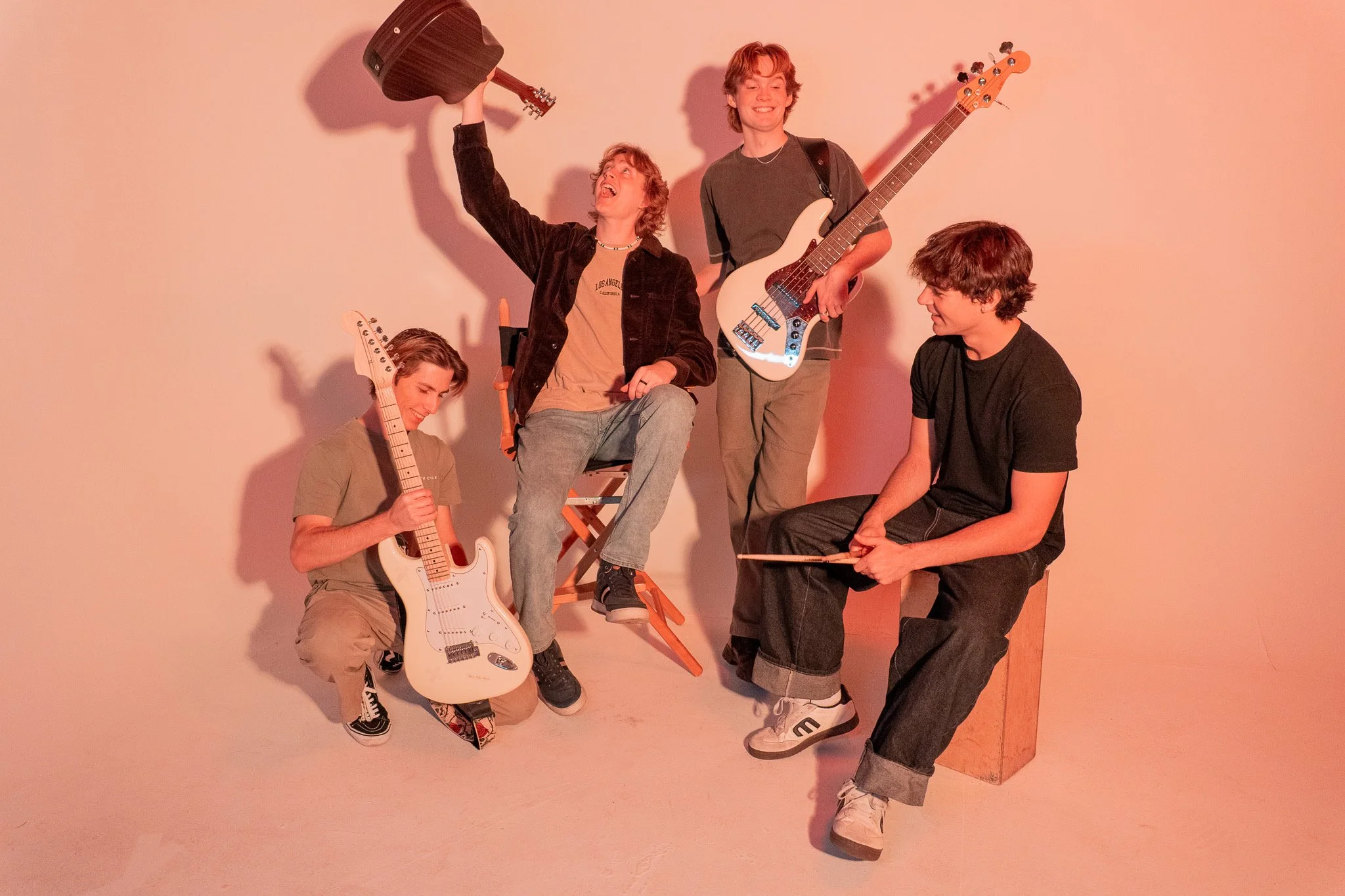 Four young men with musical instruments engaging in a lively scene against a plain background, one holding an acoustic guitar, another with a bass guitar, one sitting on a wooden box, and the fourth with a drumstick, smiling and enjoying their time t
