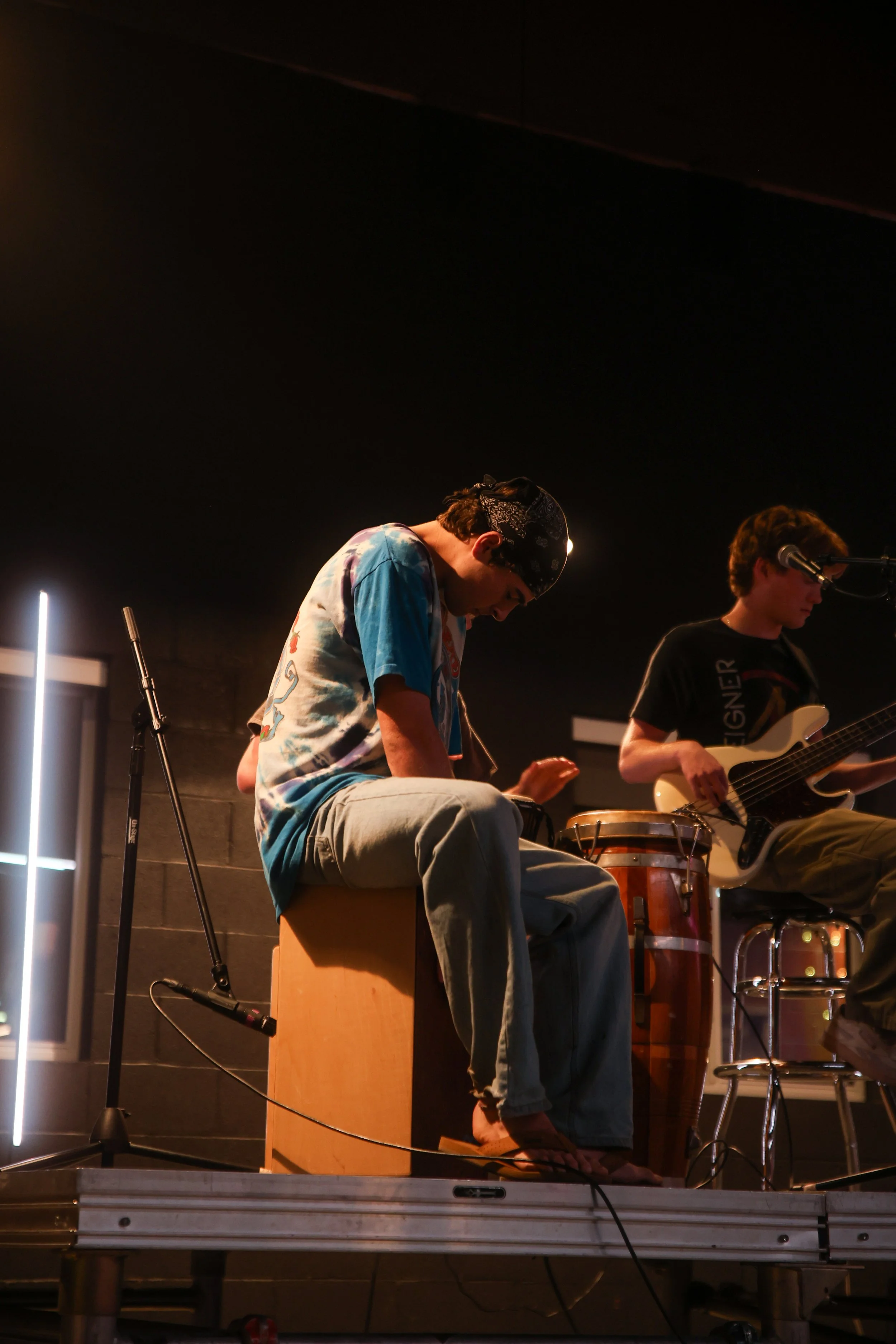 Two young musicians performing on stage with a guitar and percussion instruments, one playing the guitar and the other sitting on a cajón with a tambourine, under dim lighting.