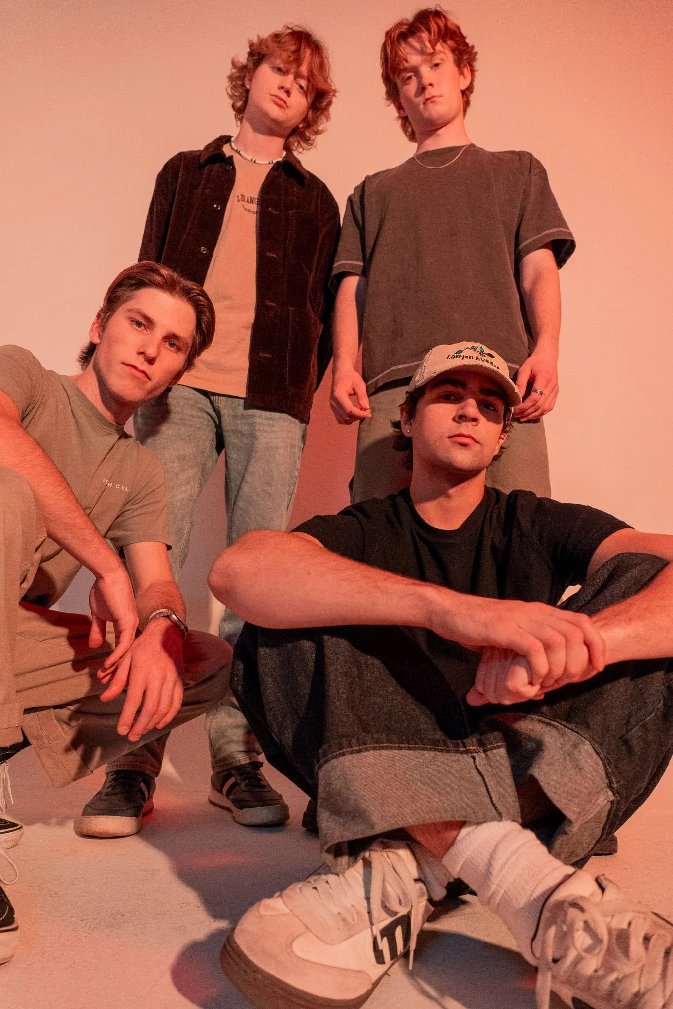 Four young men with curly or straight hair, dressed casually, pose against a plain background, with one sitting on the floor and three standing behind him.