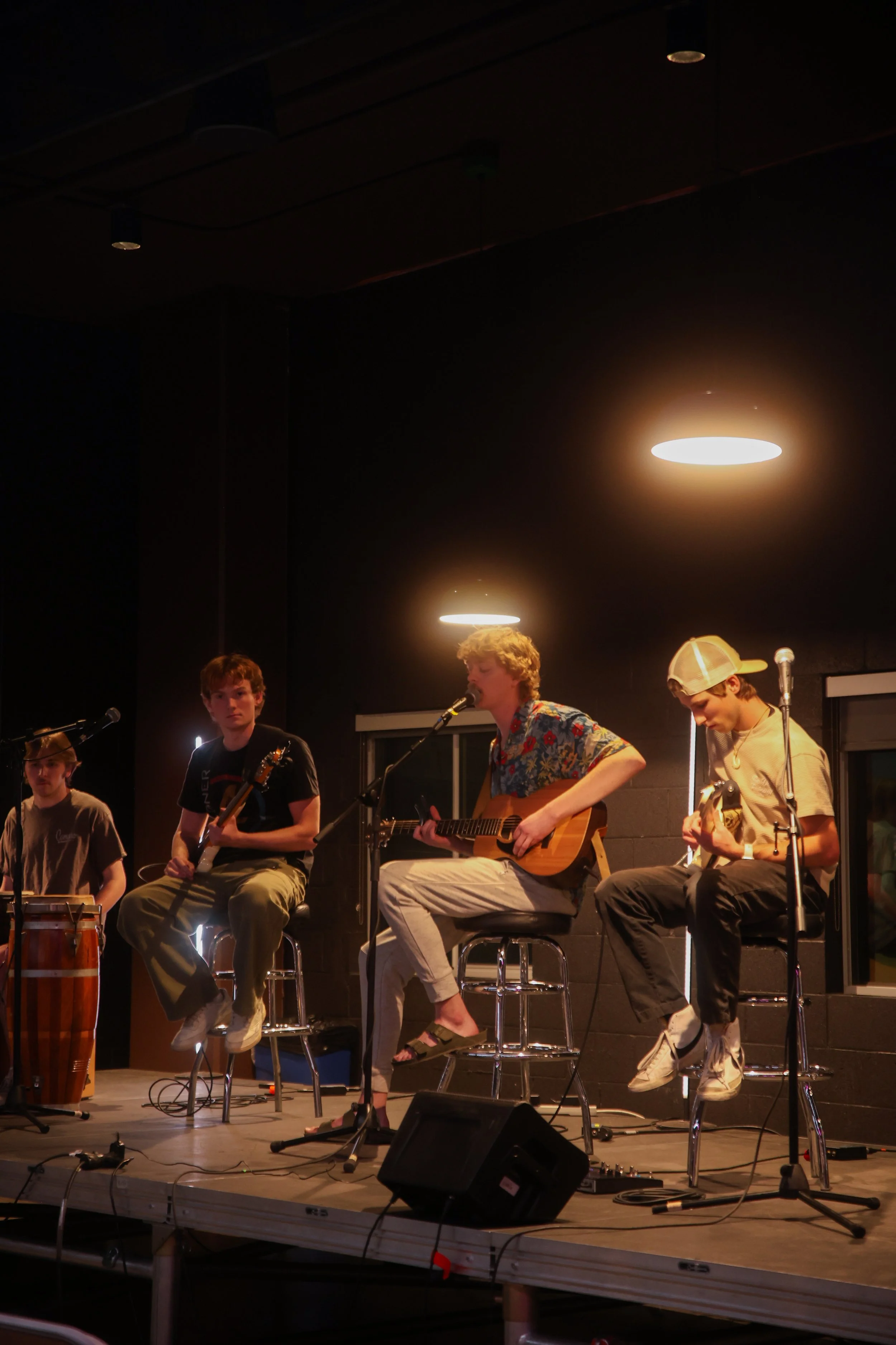 Four young males performing music on a small stage with microphones, guitars, and a drum, lit by overhead lights, with dark walls and windows behind.