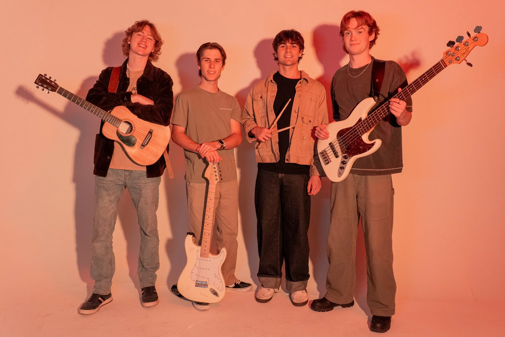 Five young men standing together, with two holding guitars, one holding drumsticks, and the others standing in front of a plain background.