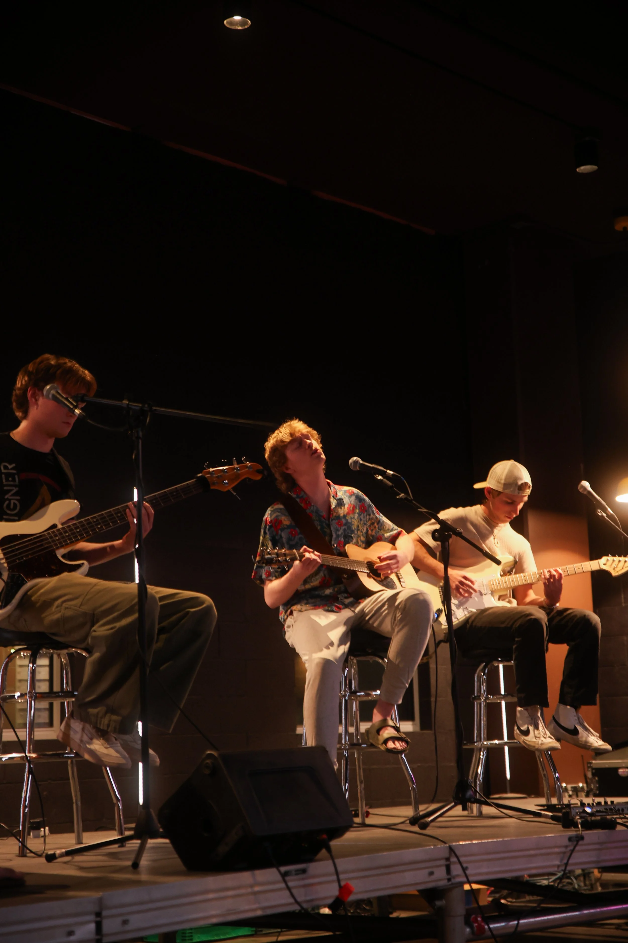 Three young men are playing acoustic and electric guitars on stage during a performance, seated on stools with microphones in front of them, with a dark background and stage lighting.
