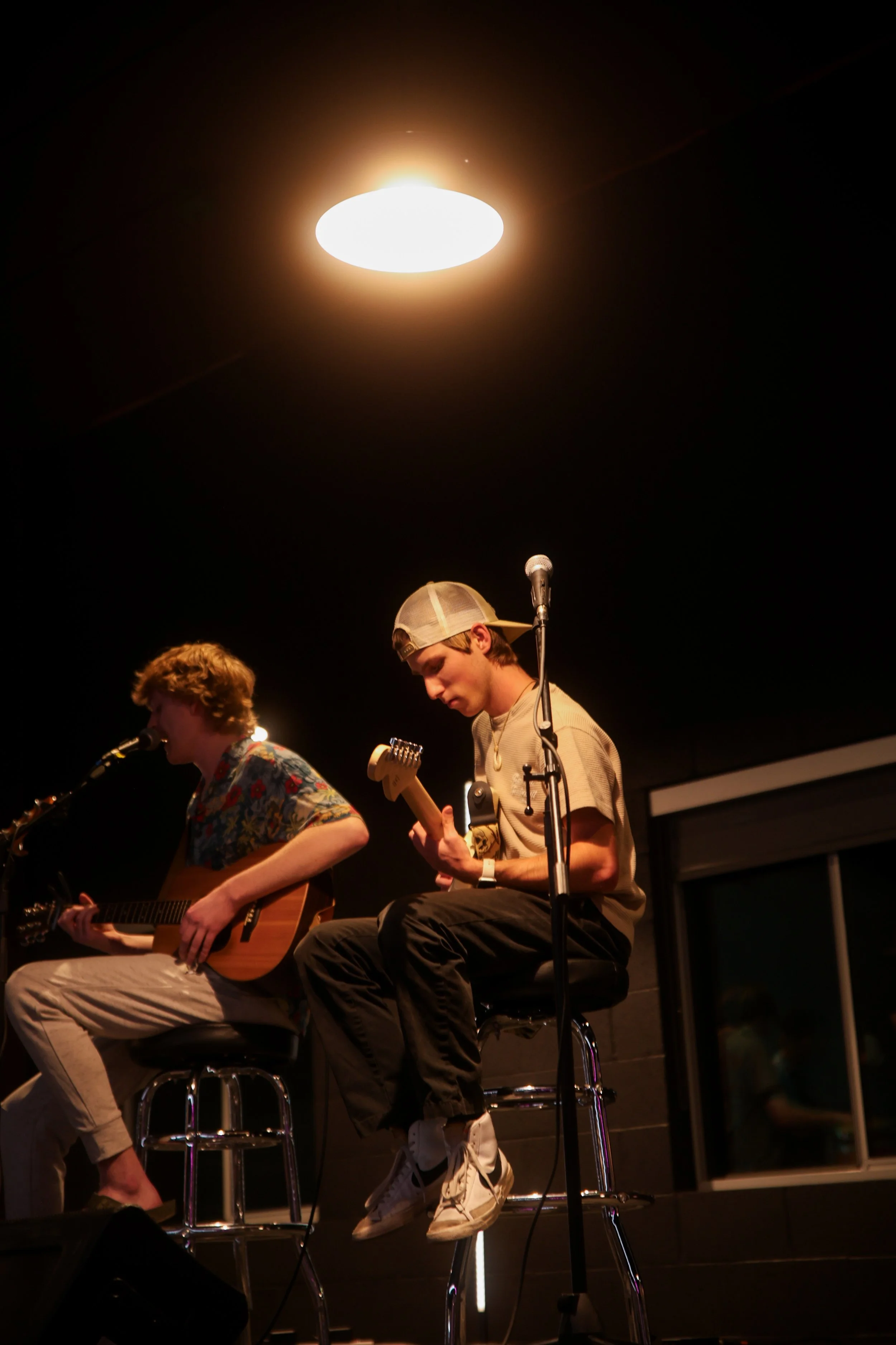 Two young men sitting on barstools, playing guitars and singing into microphones on a dimly lit stage.