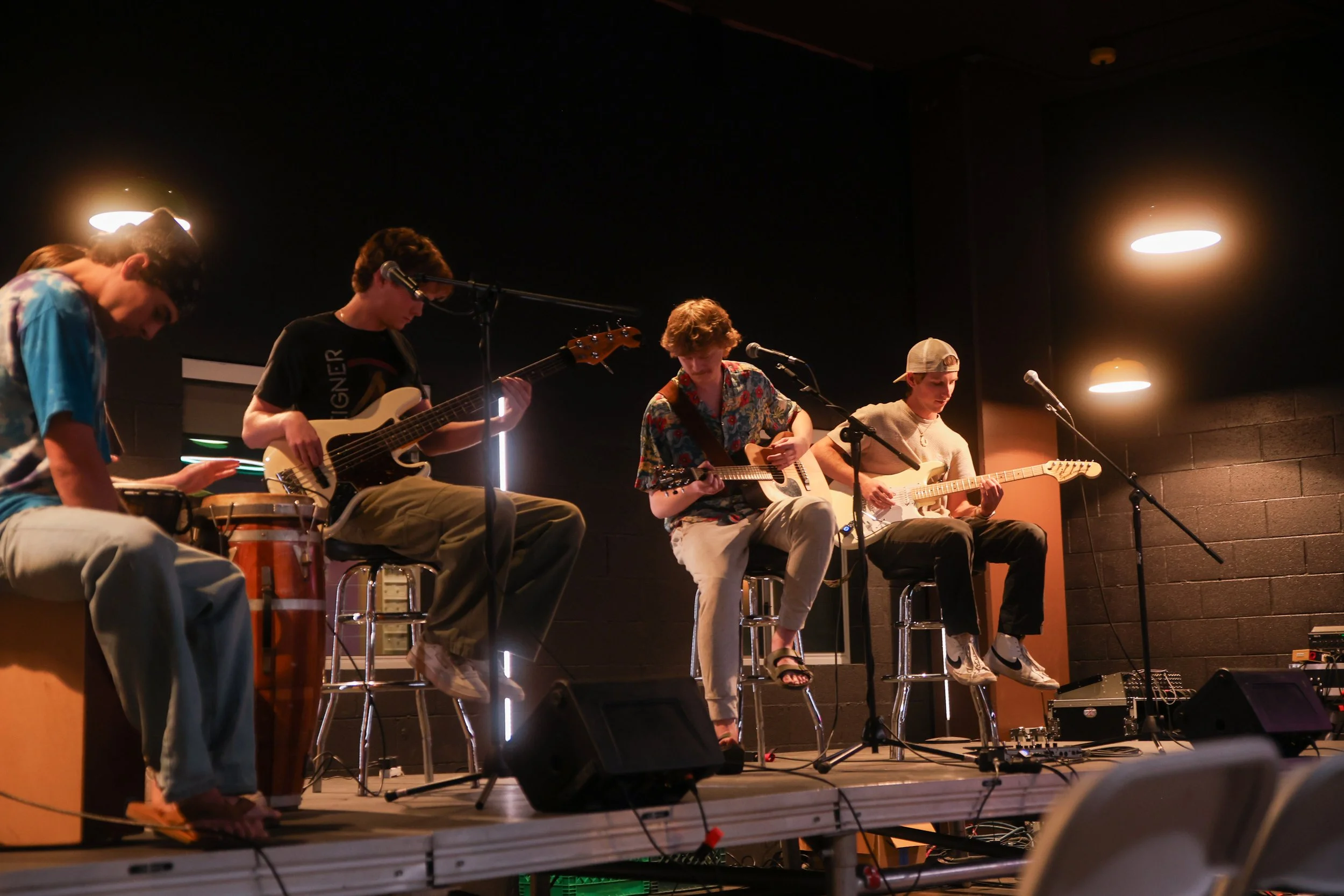 Four young men performing on stage with guitars and percussion, stage lighting in a dark indoor venue.