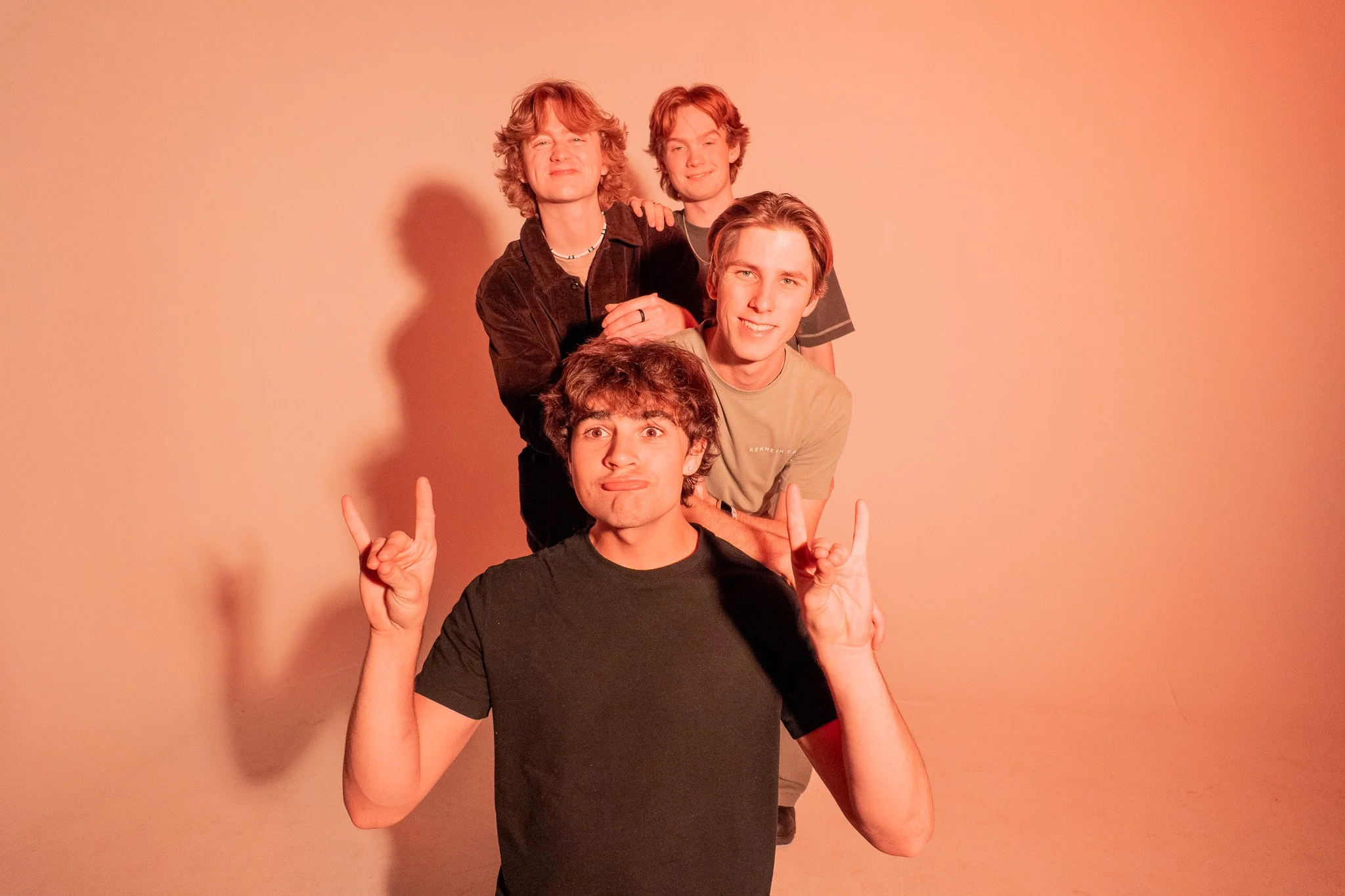 Four young men stacked on top of each other making different facial expressions and hand gestures against a plain background.