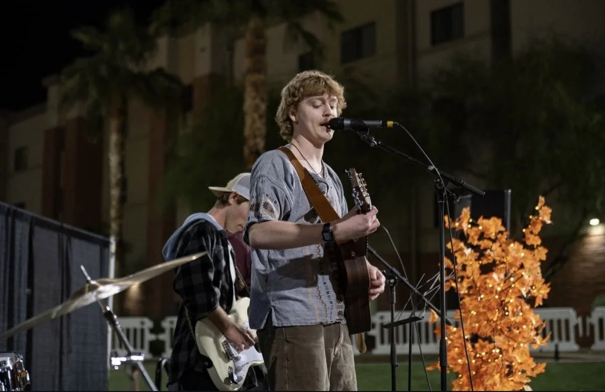 A young man with curly blond hair playing an acoustic guitar and singing into a microphone while another person plays electric guitar in the background during an outdoor night performance.