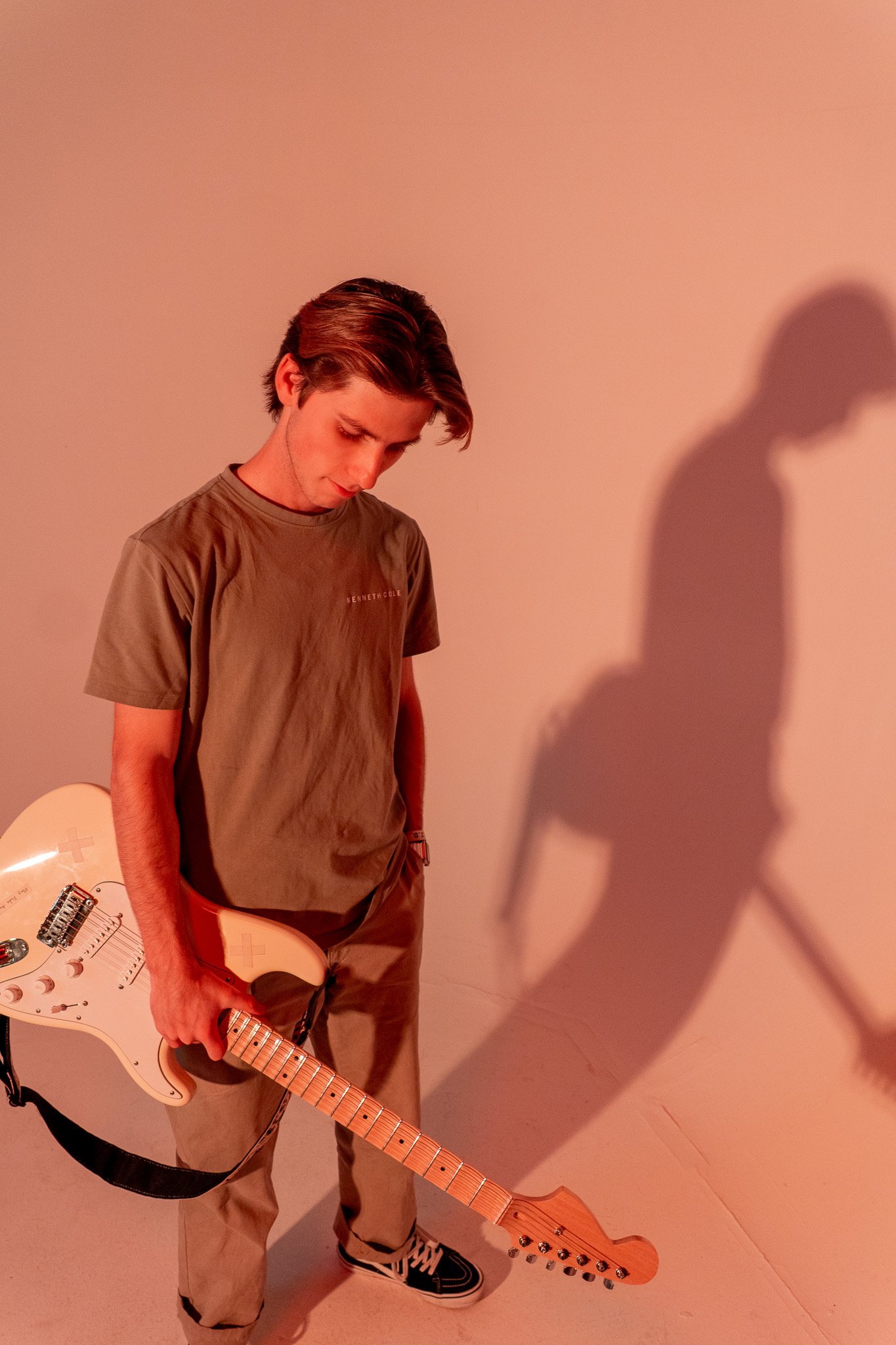Young man holding a white electric guitar standing against a beige wall with soft orange lighting, casting a large shadow of himself and the guitar on the wall.