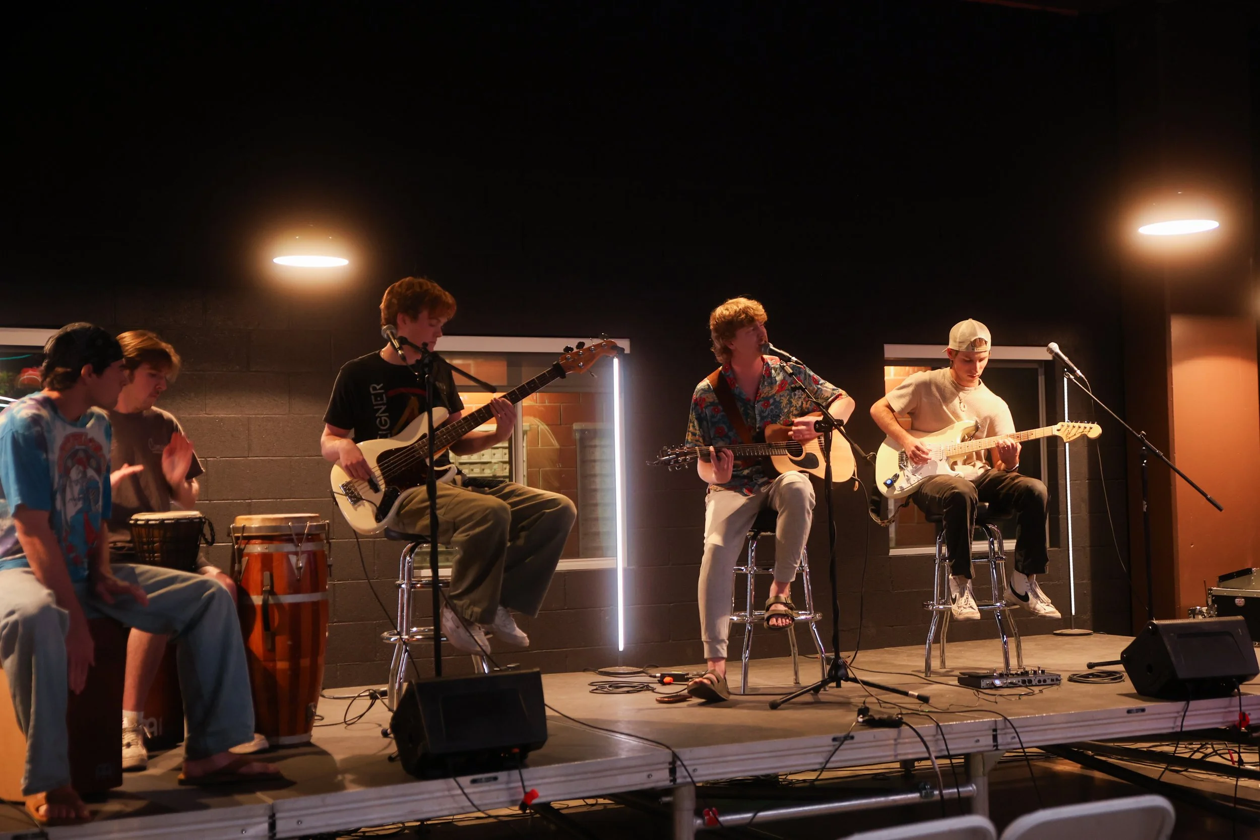 A band of five young men performing on stage with musical instruments including drums, bass, and electric guitars in a dimly lit room.