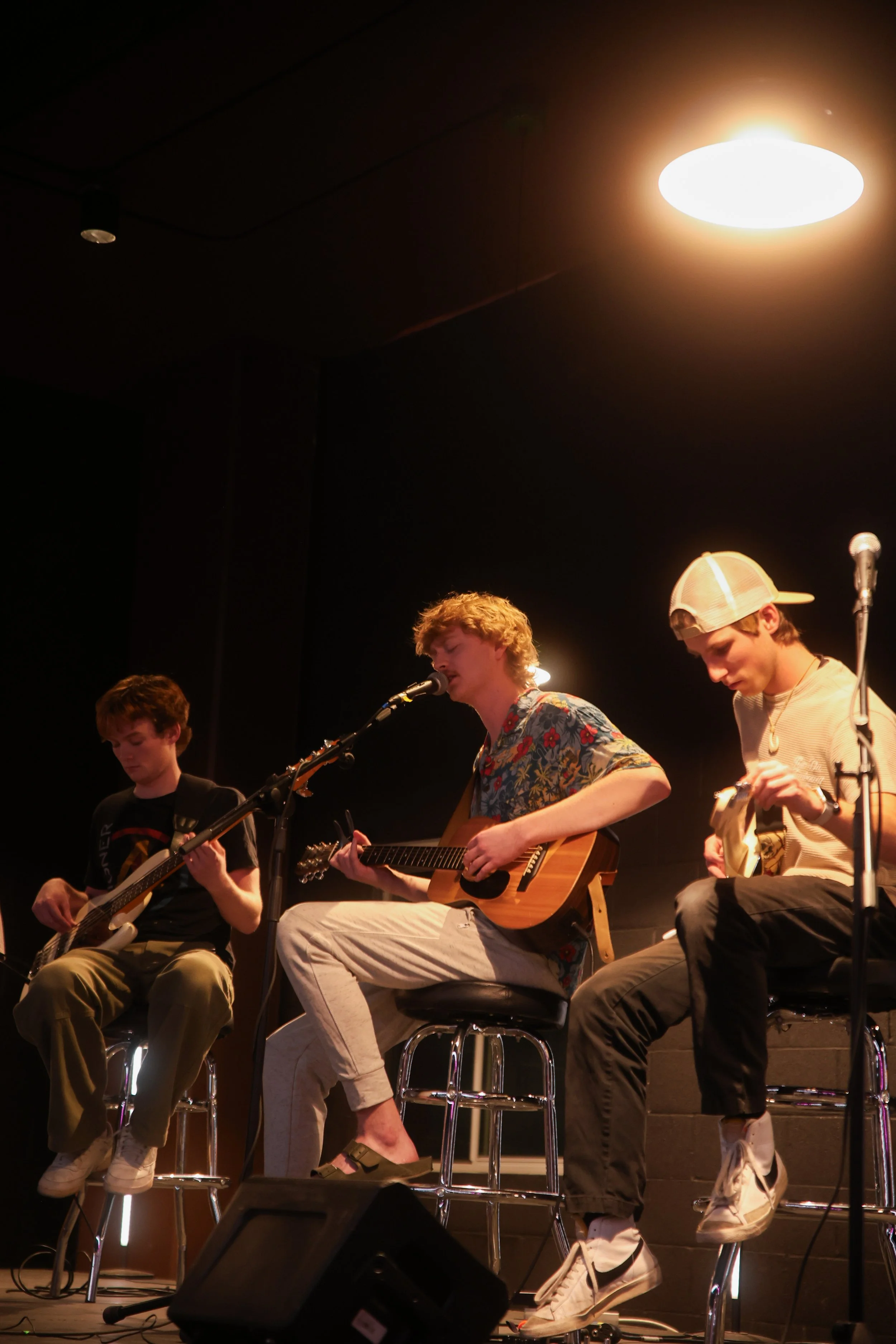 Three young men are sitting on high stools on a stage, playing guitars and singing into microphones, under warm stage lighting.